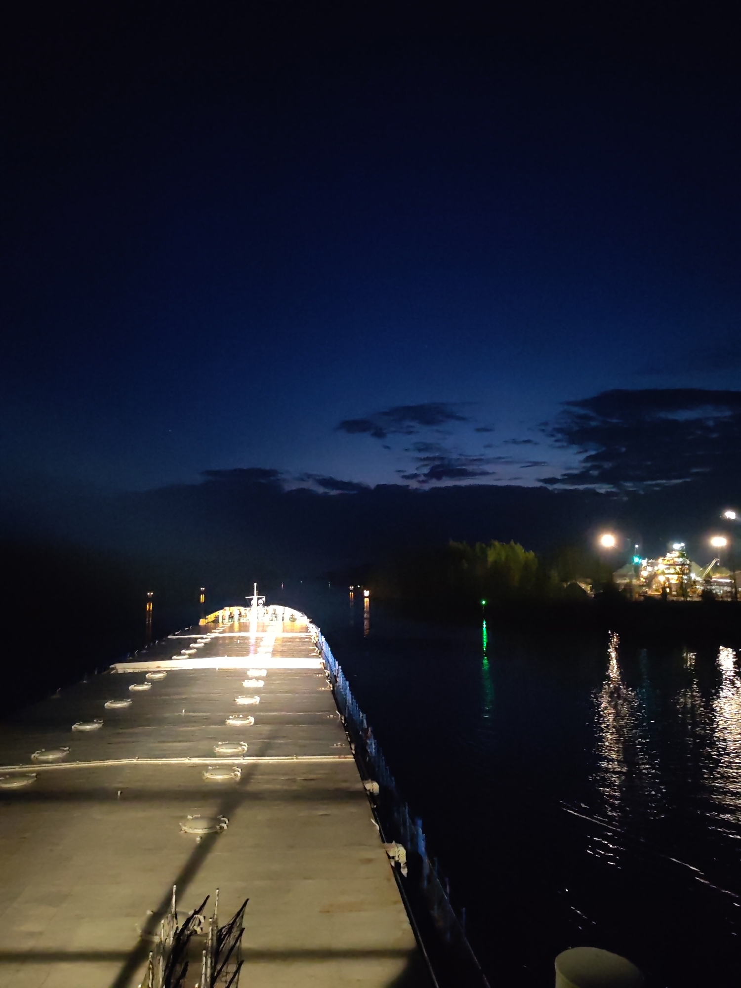 The image depicts a nighttime view from the deck of a large ship or barge. The deck is illuminated with a series of lights. The ship is traveling along a waterway. On the right side of the waterway, there is an industrial area with brightly lit buildings, storage structures, and a green light. The sky is a deep blue, with some clouds near the horizon. It could be in or near Александровское, Russia.