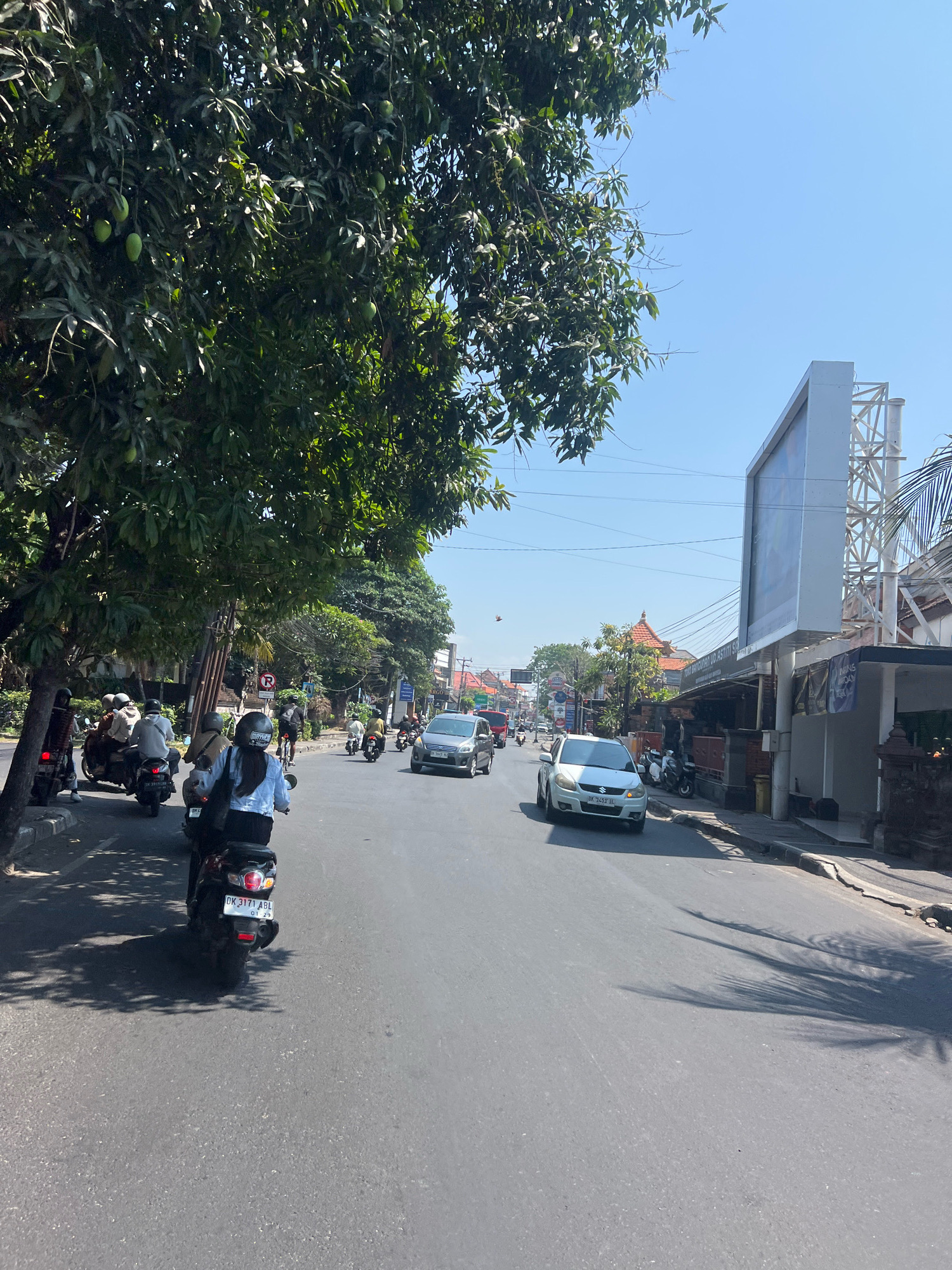 The image depicts a street scene in Denpasar, Indonesia, on a clear, sunny day. A paved road dominates the foreground, with several vehicles traveling along it. On the left side of the road, a large tree with dense green foliage provides shade. Several motorcycles and a few cars are visible. One motorcycle in the foreground has a rider wearing a white shirt and a black helmet. Further down the road, a silver car is turning.
The right side of the road features buildings, including a structure with a large white billboard above it. Various shops and smaller buildings line the street. Signs are visible on some of the buildings, though not all are legible. The sky is a clear, bright blue, indicating good weather. The overall impression is a busy urban environment with typical Indonesian street traffic.