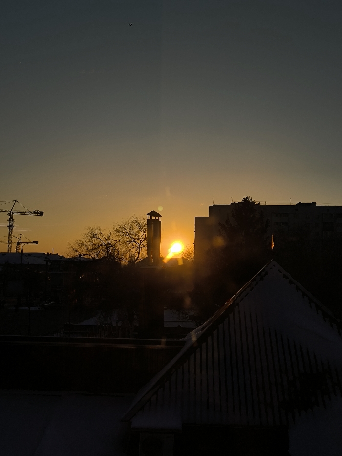 The image depicts an urban landscape in Vinnytsia, Ukraine, during either sunrise or sunset, characterized by strong backlighting. The prominent central structure, identified as the Vinnytsia Water Tower (Mykhaila Kotsiubynskoho St, 30), is silhouetted against the low sun.
The sky occupies the upper two-thirds of the frame, transitioning from a dark grey-blue at the zenith to a vibrant orange and yellow at the horizon, where the sun is a bright, intense orb partially obscured by foreground elements and causing significant lens flare. A small, dark silhouette of a bird is visible in flight in the upper-left quadrant of the sky.
In the foreground, occupying the lower-right, is the dark, snow-dusted silhouette of a steeply pitched gable roof with visible vertical siding or planks. A small, light-colored flag-like ornament is positioned at the roof's peak.
The midground and background consist primarily of dark silhouettes. To the left, one or more tall construction cranes are visible, indicative of ongoing development. Interspersed across the middle ground are bare deciduous trees, suggesting a winter season, with some visible snow accumulation on lower structures. To the right of the water tower, several multi-story buildings, consistent with residential or commercial structures, form part of the city skyline. No individuals or specific actions are discernible within the frame. The overall environment suggests an urban area under cold, clear sky conditions.