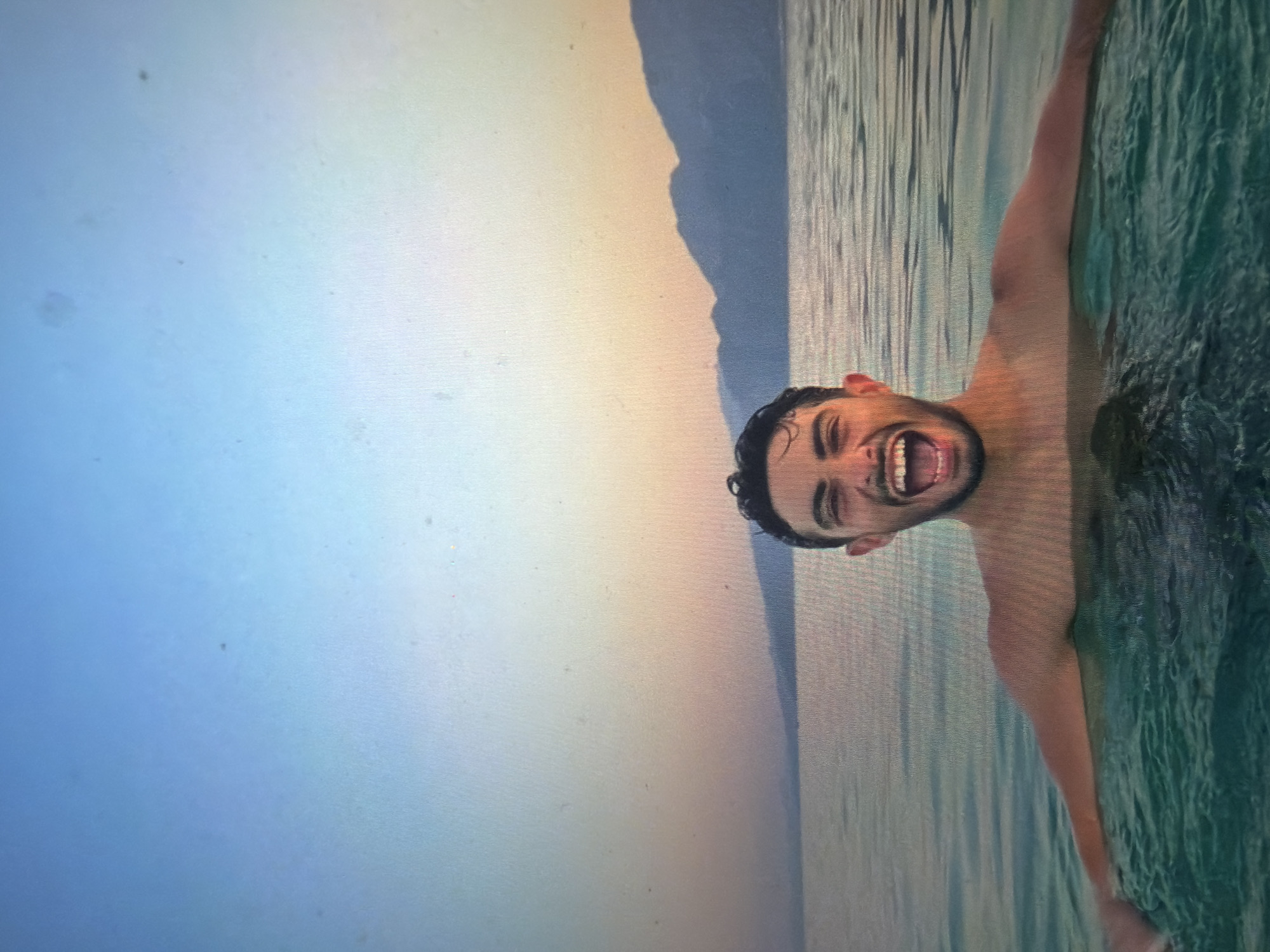A young adult male is depicted chest-deep in a body of water, facing forward with a wide-open mouth, exposing his teeth, and wide-open eyes. His arms are partially submerged and extended slightly outwards. The water in the foreground is a dark greenish-blue with visible ripples, transitioning to a lighter, reflective grey-blue towards the horizon. In the mid-ground, a broad body of water extends to meet a range of distant, dark mountainous landforms silhouetted against the sky. The sky above exhibits a color gradient, shifting from a light blue at the top of the frame to shades of orange, yellow, and pink closer to the horizon, consistent with either a sunrise or sunset. The image displays digital artifacts, including a visible grid pattern and numerous small specks and smudges, particularly in the sky area.