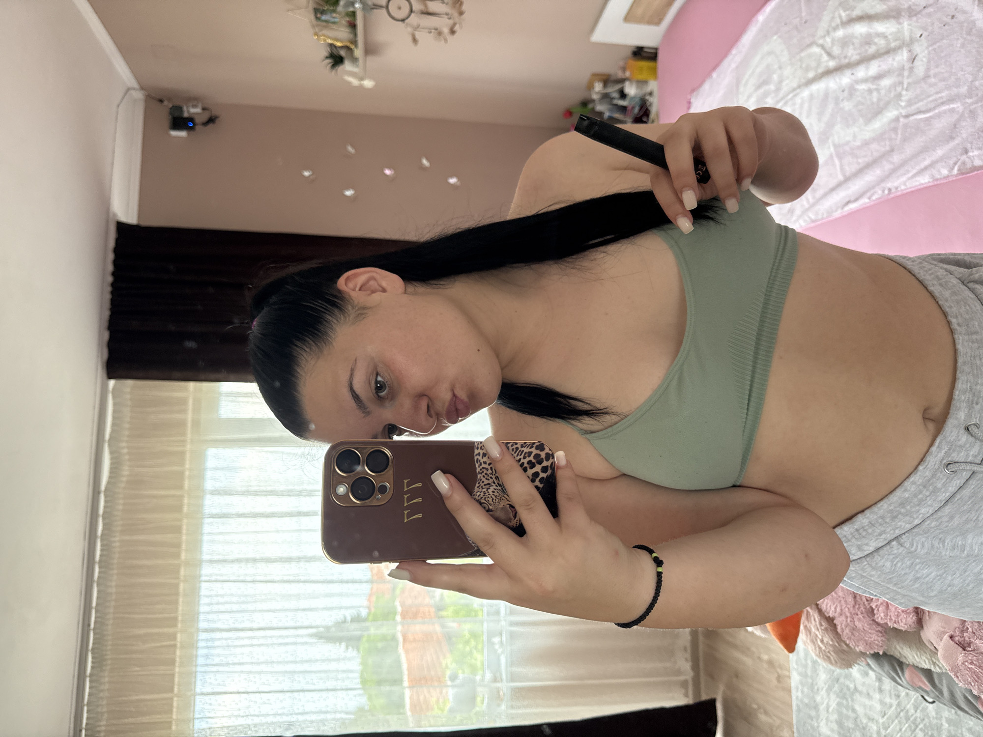 A young woman takes a selfie in a mirror. She is wearing a light green sports bra and grey sweatpants. Her long, dark hair is pulled back. She is holding a black electronic device in her right hand and her iPhone in her left. The phone case is brown with a leopard print accent and three gold initials that appear to spell "FFF." Her nails are painted a light beige color. She is puckering her lips as she looks into the mirror.
The background suggests a bedroom. There is a bed with pink bedding and a white comforter visible behind her. The walls are a light beige color. A window with sheer white curtains is to her left, showing greenery outside. The image has a vertical orientation and is taken in natural light, likely during the daytime. The overall mood appears casual and relaxed. There is a small black device with a blue light mounted on the wall in the upper left portion of the mirror's reflection.