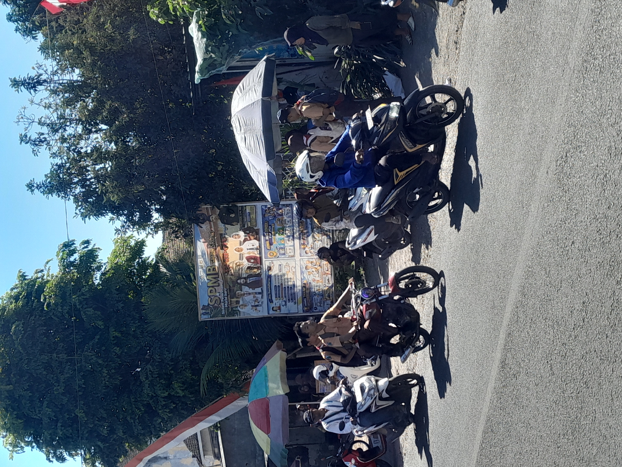 The image depicts a street scene in Srono, Indonesia, on a bright, sunny day. Several people are on motorcycles, navigating the asphalt road. In the foreground, a person in a blue jacket and white helmet rides a black and yellow scooter. To their left, a person wearing a white shirt rides a white scooter. Behind them, a young person in a brown school uniform rides a red motorcycle, with another person in a similar uniform on the back. Further back, under a white umbrella, a group of people are gathered, some appearing to be students in uniform. A large banner with text and images, possibly an advertisement or informational poster, is visible behind them. Trees and buildings line the street, with a clear blue sky above. The atmosphere suggests a normal day with people commuting and congregating.