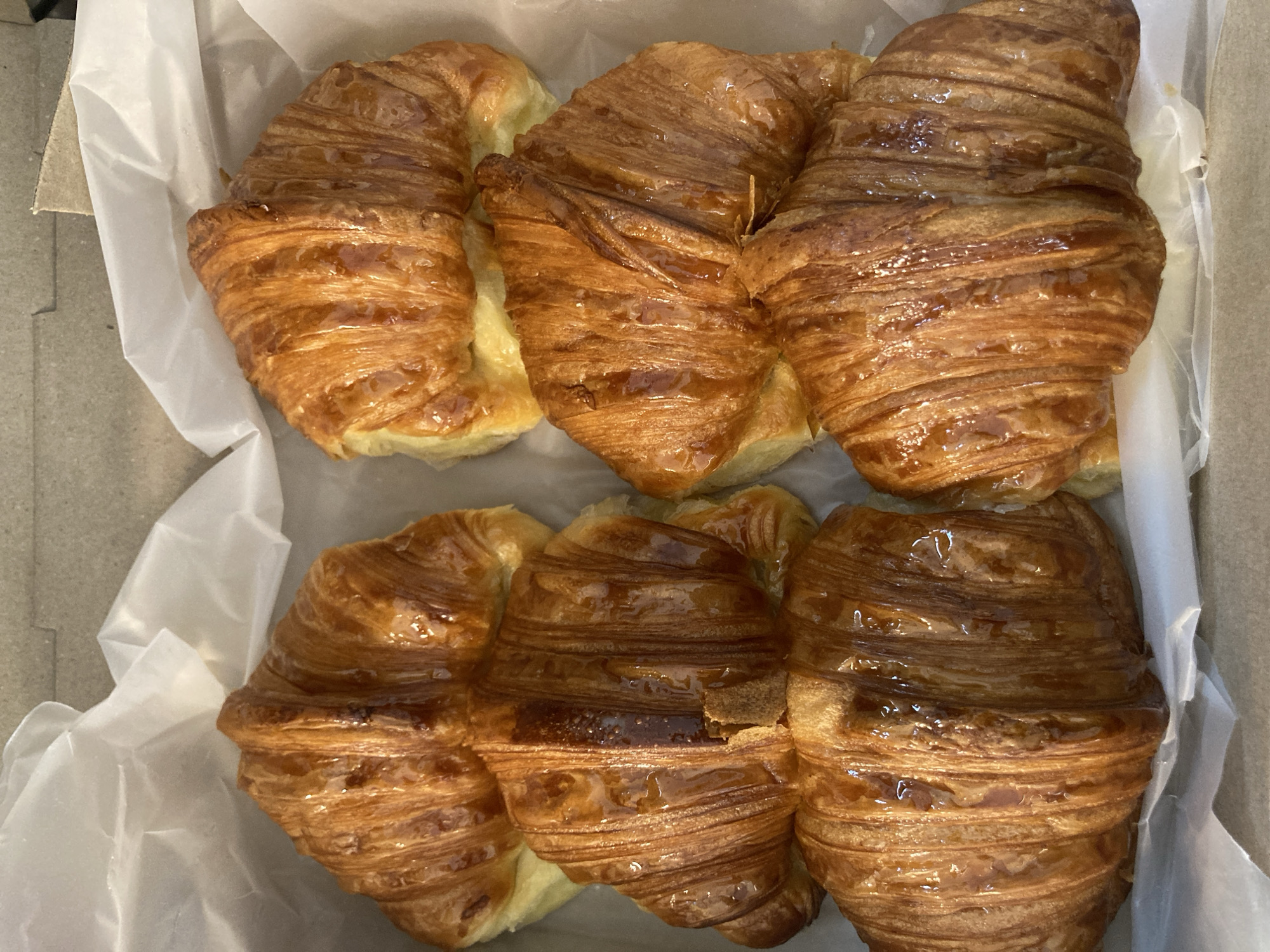 The image shows six croissants arranged in two rows of three within a container lined with white tissue paper. The container itself appears to be made of cardboard. The croissants are golden brown, displaying a layered and flaky exterior, suggesting they have been freshly baked. The lighting in the image highlights the glaze on the croissants, creating a shiny surface. The scene takes place in a location whose city and district are Unknown.