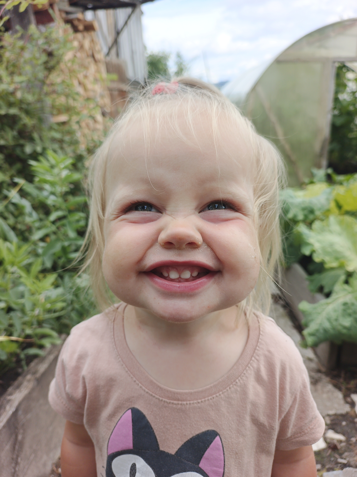 In Medvedyovka, Russia, a young Caucasian child with blonde hair, tied up with a pink elastic band, is pictured in a close-up shot. The child is wearing a light pink t-shirt with a black cat design featuring pink ears and white eyes. The child has rosy cheeks and is smiling broadly, showing teeth, with their eyes looking to the side and partially closed. The background reveals a rural setting with green foliage, a wooden structure, and a greenhouse. The lighting suggests it is daytime.