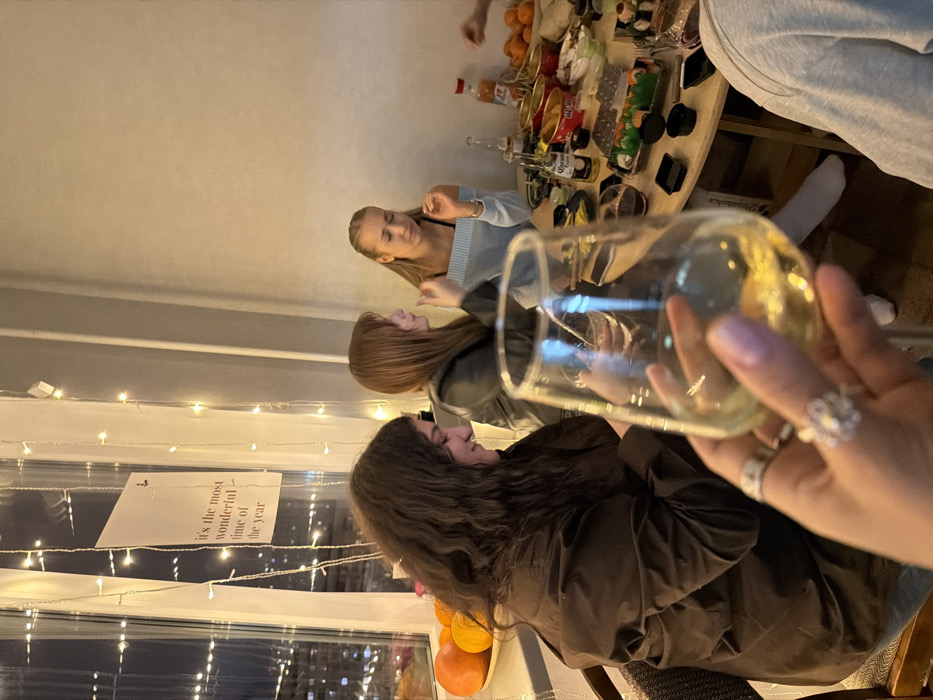 The image captures an indoor social gathering taking place during the evening or night in an apartment located in Moscow, Russia. The room is softly lit by artificial light and decorative string lights cascading vertically along a large window overlooking a city skyline dotted with lights.
In the immediate foreground, a person's hand, adorned with light pink nail polish, holds a stemmed glass filled with a pale yellow liquid, indicative of white wine. Two rings are visible on the fingers: a simple band on the ring finger and a prominent, possibly floral or star-shaped, ring on the middle finger.
Around a light-colored, possibly circular, table, at least four young women are seated. To the left, a woman with long, dark, wavy hair, wearing a dark brown jacket, is observed looking downwards with a faint smile. Adjacent to her, a woman with shoulder-length reddish-brown hair, dressed in a black jacket, is seen in profile, appearing to be speaking or gesturing. Further to the right, a blonde woman with her hair pulled back, wearing a light blue off-the-shoulder top, is looking down, seemingly engaged in the activity at the table. A fourth individual, wearing a light grey long-sleeved top, is partially visible in the bottom right corner.
The table is densely covered with various items: multiple bottles of Corona beer, a bottle with an orange label, several red and gold containers filled with snacks, black plastic trays holding sushi, and at least one other glass containing a dark beverage. A bowl of oranges or tangerines is situated on a shelf by the window. A white rectangular sign, bearing the English phrase "it's the most wonderful time of the year," is affixed to the window frame amidst the string lights. The walls of the room are plain and light-colored.