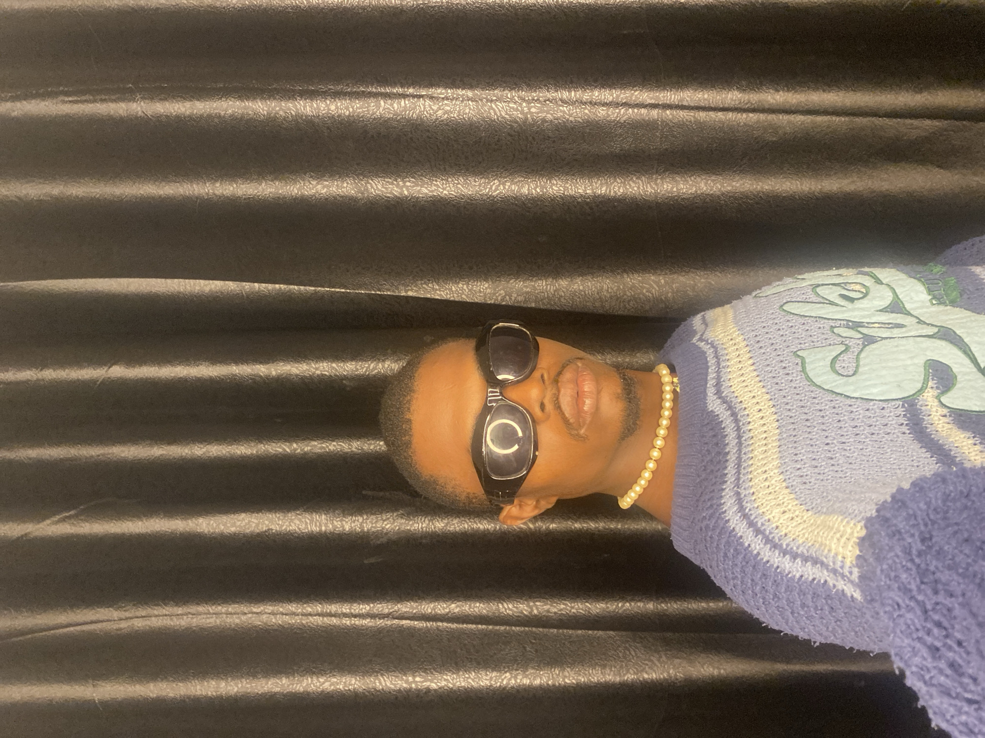 In Abakaliki, Nigeria, a young Black male stands centered in the frame, facing the camera directly. He wears dark, wraparound sunglasses that cover his eyes and bridge of his nose, and a pearl necklace. His lips are slightly pursed, and he has a mustache and goatee. A blue and white striped sweater with a green graphic design on the chest covers his upper body. The background consists of dark, textured curtains, possibly made of satin or a similar shiny material, with vertical folds that create a ribbed effect. The lighting appears to be coming from the front, casting subtle shadows.