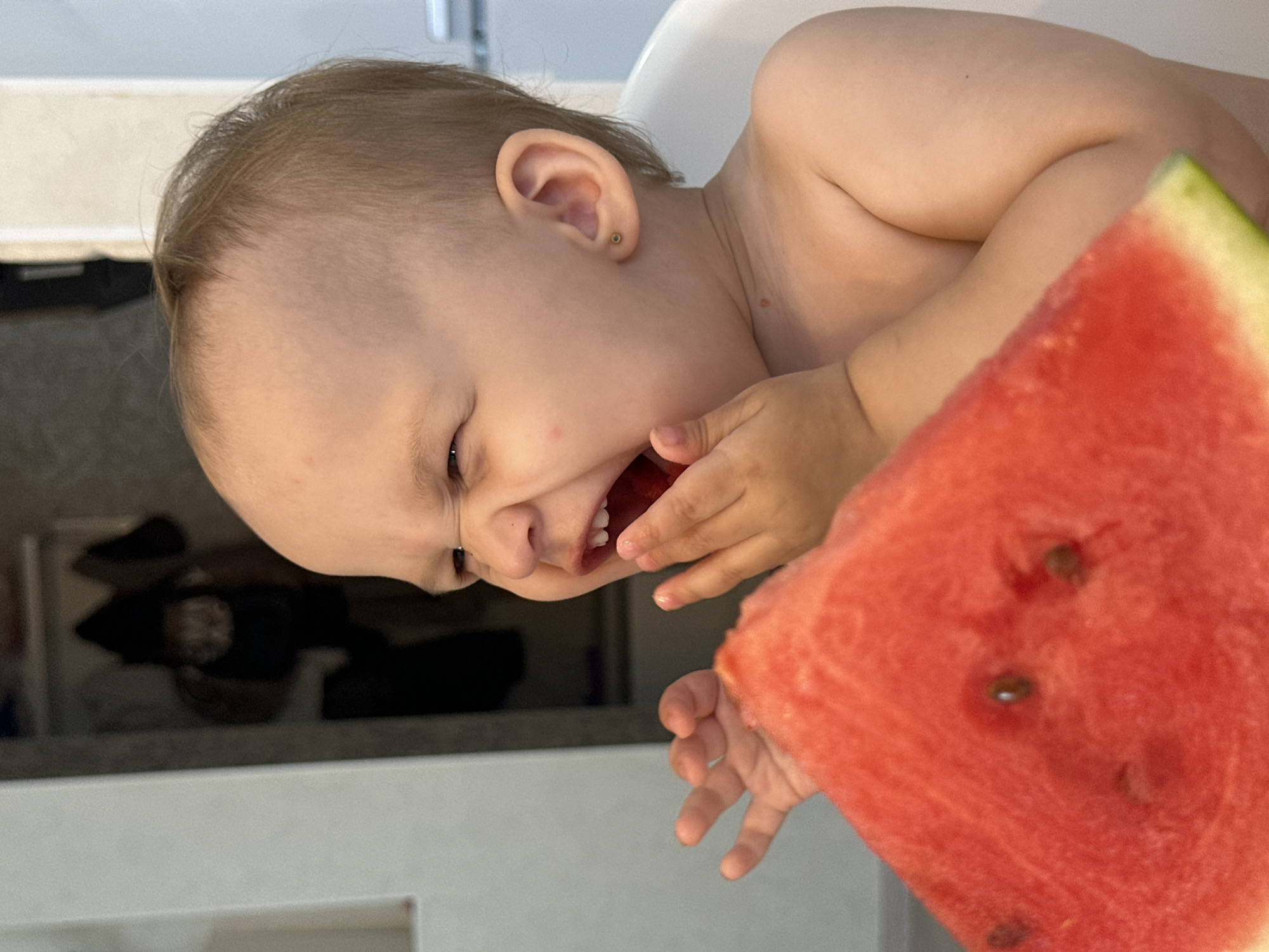 A close-up, vertical shot shows a young child being fed a slice of watermelon. The child is leaning forward, mouth open, eyes squeezed shut as they bite into the fruit. Their right hand is reaching towards the watermelon, while their left hand is obscured. The watermelon is a vibrant red with visible black seeds. The child has fair hair and is wearing a small gold stud earring in their left ear. The background is blurred, with hints of a kitchen environment, including countertops and cabinetry. The lighting suggests it is daytime. The child's expression conveys intense focus and enjoyment of the watermelon. The scene is likely indoors, possibly in a home kitchen or dining area, in Chaikovsky, Russia.