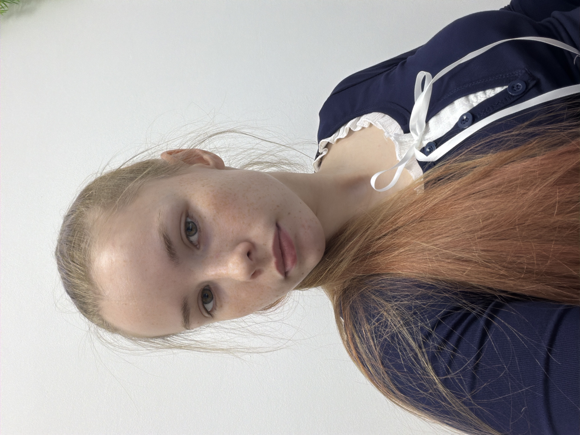 The image shows a young woman with fair skin and freckles. Her hair is light blonde at the roots and transitions to a reddish-brown towards the ends. She is wearing a dark blue top with white trim and a white ribbon tied at the collar. The background is a plain white surface. There is no information in the image indicating a location in Berezniki, Russia.