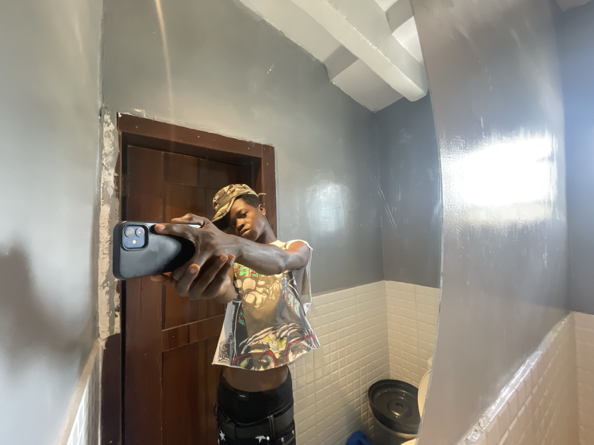 A young, dark-skinned male stands in a bathroom, positioned to the left of the frame, and is holding a smartphone, pointed to the left. He wears a camouflage baseball cap and a sleeveless t-shirt. The room is painted gray above a white tiled wainscotting. A dark wooden door is behind him to his left. A trash can is partially visible in the lower right corner. Location information is not available in this image.