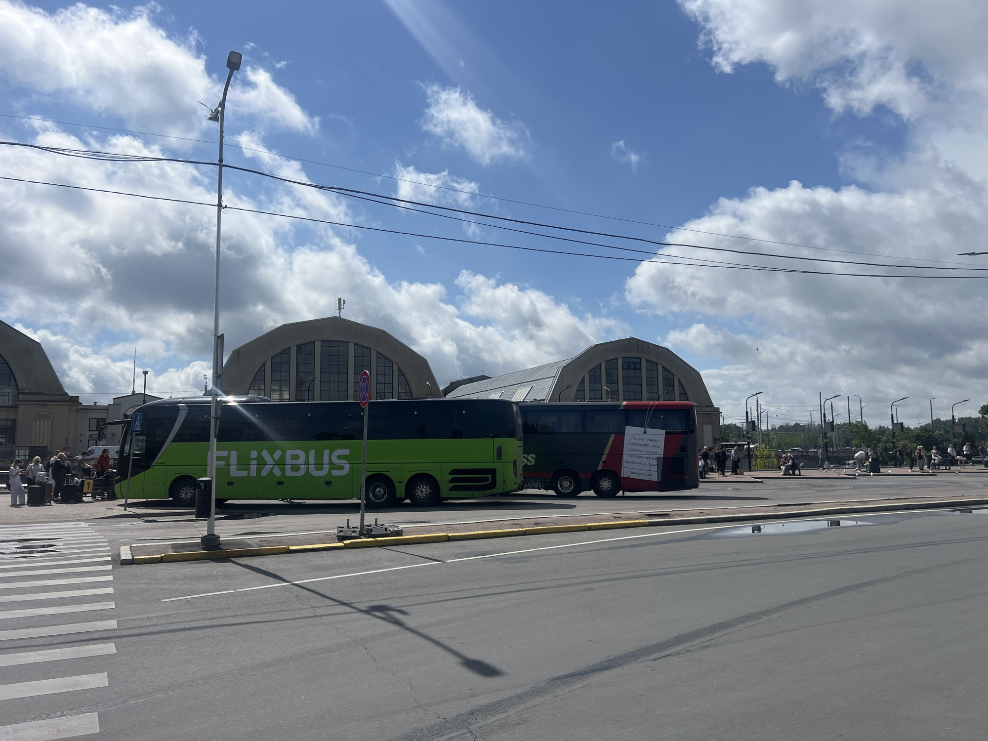 This image depicts a scene at the Riga Central Bus Station in Riga, Latvia. Two intercity buses are parked on the asphalt. The bus on the left is green and white with "FLIXBUS" emblazoned on its side. The bus on the right is red and black with text on its side indicating destinations such as "TALLINN • PÄRNU" and "RIGA." Several people are visible near the buses, some with luggage, suggesting they are passengers. In the background, a large, arched building, characteristic of the bus station's architecture, is visible. The sky is partly cloudy, and a street light is prominent in the foreground, casting a shadow on the road. A crosswalk with white stripes is also visible in the lower left portion of the image.