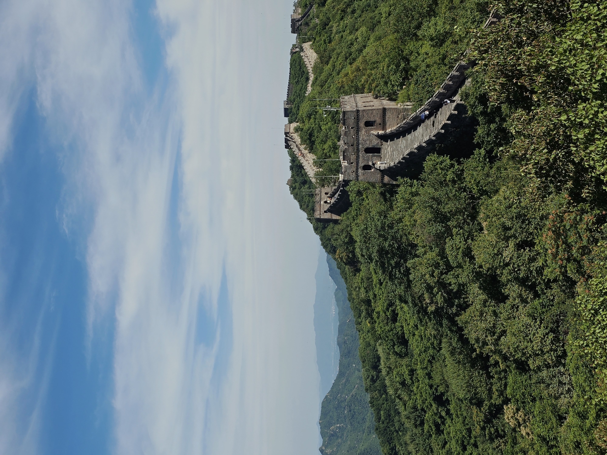This is a daytime, eye-level shot of the Great Wall of China, specifically at the Mutianyu section in Huairou District, China. The ancient stone fortification winds along a lush, densely forested mountain ridge under a bright blue sky with scattered white clouds. Watchtowers are visible at intervals along the wall, with several tourists walking along the paved pathway. The wall itself is constructed from stone and brick, with crenellated battlements and arched doorways in the towers. The surrounding environment is characterized by rolling green hills covered in a thick canopy of trees, creating a vibrant natural landscape that contrasts with the man-made structure. In the distance, hazy mountain ranges are visible.