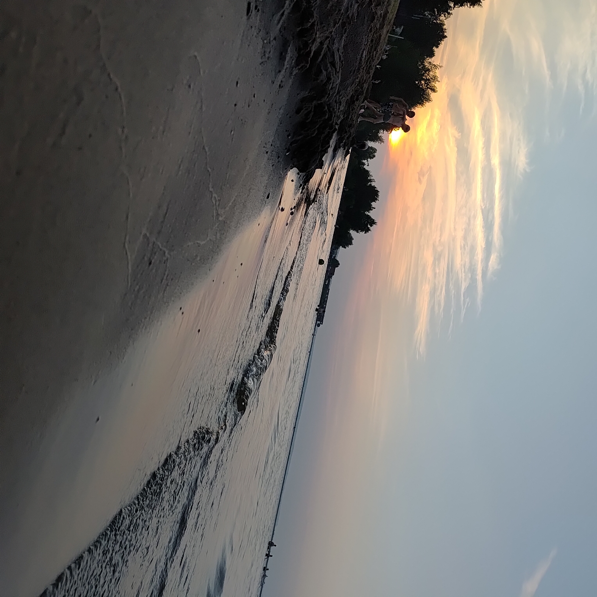 The image depicts a sunset scene at a beach, likely on the Kama River, in Chaikovsky, Russia. In the foreground, a sandy beach slopes down towards the water. Two young males are visible on the sandy embankment, one behind the other, wearing swimwear. The sun is setting on the horizon, casting a warm orange and yellow glow across the sky, with wispy clouds. The water is calm with gentle waves lapping at the shore. In the background, trees line the distant bank of the river, and a long structure, possibly a pier or building, is visible along the water's edge. Other individuals can be seen scattered along the beach and in the water, enjoying the evening. The overall atmosphere is serene and recreational.