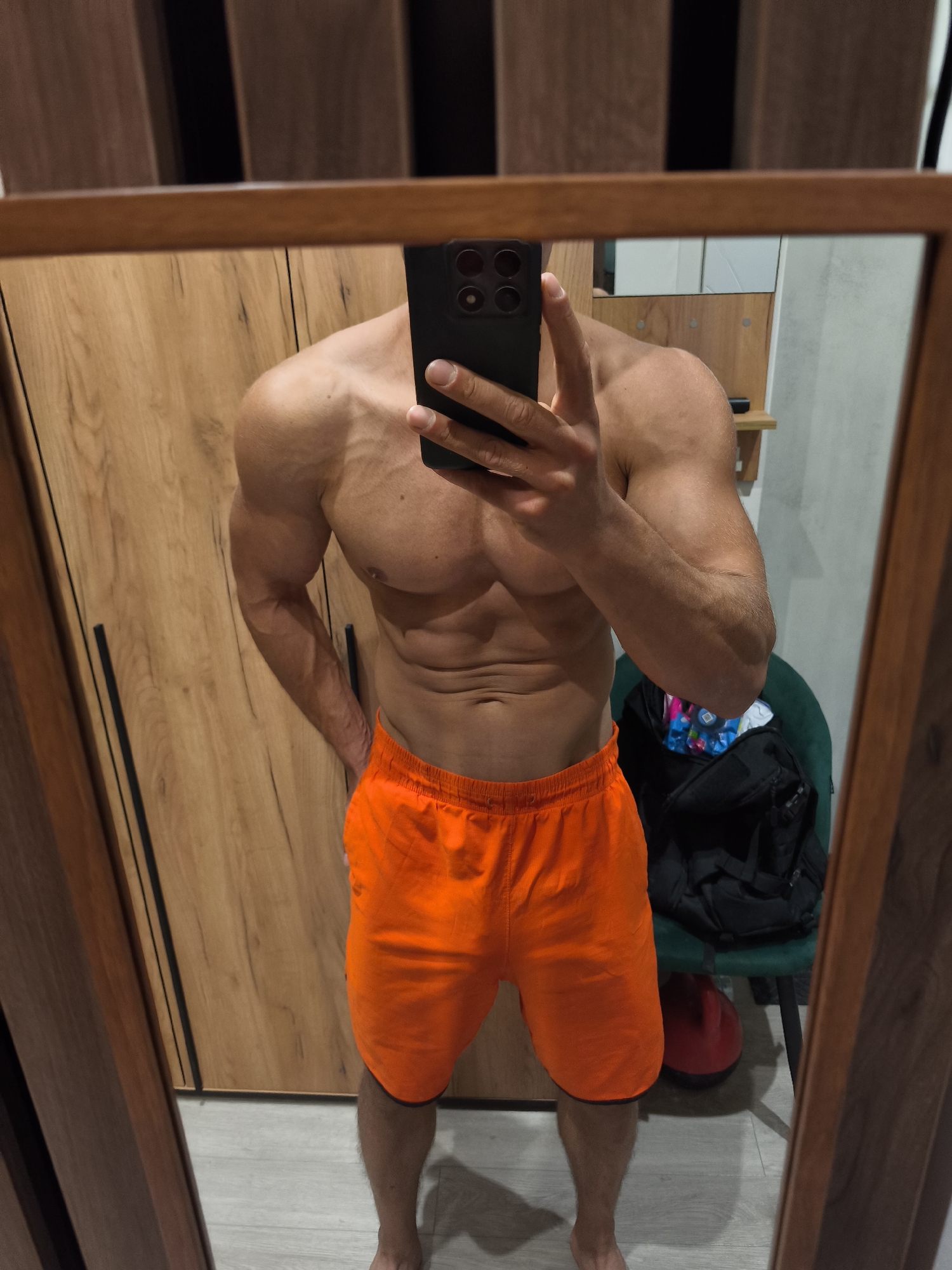A man is taking a mirror selfie in what appears to be a bedroom or dressing room. He is shirtless, wearing orange shorts, and has a muscular physique. His phone, held up to take the photo, is visible in the reflection. The background shows a wooden wardrobe and a chair with a bag on it. There is no indication of the location being Moscow, Russia.