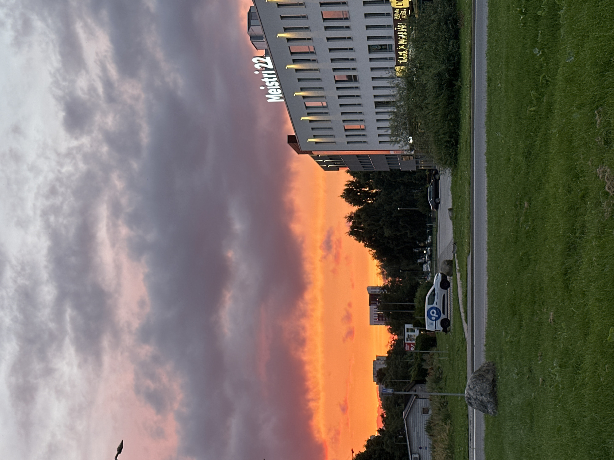 The image depicts a city scene at sunset in Tallinn, Estonia. The sky is a vibrant mix of orange, pink, and purple hues, indicating a dramatic twilight. In the foreground, a well-maintained green lawn is visible, with a large grey boulder to the left and a paved road with a white van parked on the right side of the frame. The van has a blue circular logo on its side.
Dominating the right side of the image is a modern, multi-story building with the illuminated signage "Meistri 22" on its roof. The building features a facade of white panels and numerous rectangular windows, with vertical lights illuminating sections of the exterior. Below the building, illuminated signage for businesses can be seen, including "SALAD" and what appears to be "PAGARIÄRI". Trees and other smaller buildings are present in the background, interspersed with glimpses of the city skyline. The overall impression is one of an urban environment transitioning from day to night under a colorful sky.