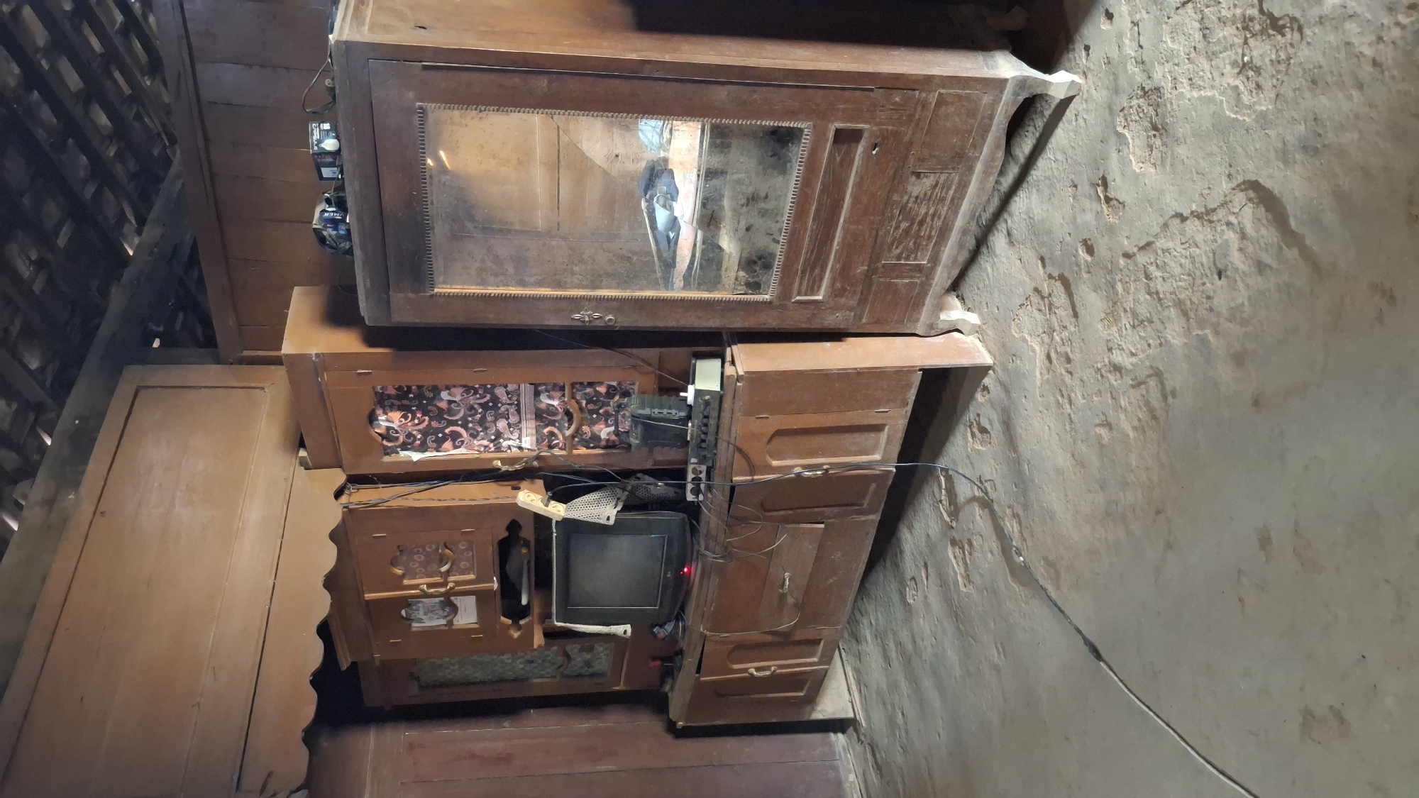 This is a medium shot, eye-level, full-color photograph of a room with wooden furniture in Pucakwangi, Indonesia. The room appears to be a living area or bedroom. The primary focal point is a large, dark wood entertainment center unit filled with various objects, including a small television with a red light on, a radio, speakers, and several cables. To the right of the entertainment center is a tall, dark wood cabinet with a glass door, revealing items inside. The room has a rustic feel with a wooden ceiling and exposed wooden beams. The floor is made of concrete with some uneven patches. The lighting is dim, suggesting it might be indoors during the day or at night with artificial lighting. There are no people visible in the image.