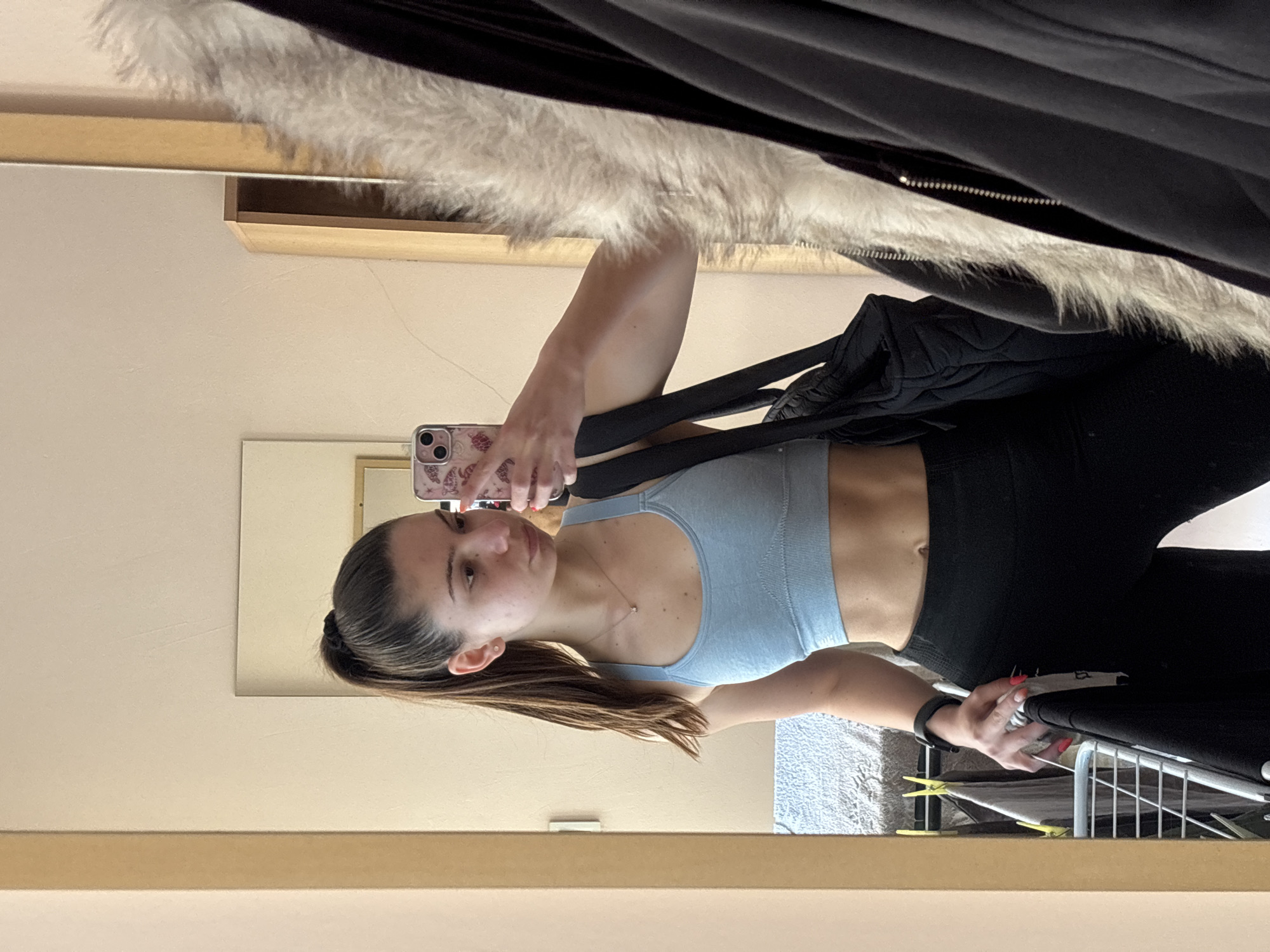 An indoor, mirror selfie captures a young woman from the waist up. She appears to be in her late teens or early twenties with light skin and dark brown hair, pulled back in a ponytail or bun, with some hair falling forward. She is wearing a light blue, possibly ribbed, sports bra that reveals her toned midriff, and black leggings. A simple necklace with a small pendant is visible around her neck, and she wears a black fitness tracker or watch on her left wrist. Her fingernails are painted a reddish-orange hue.
She is holding a smartphone with a distinctive pinkish-red case featuring a butterfly or floral pattern in her right hand, capturing her reflection. Her expression is neutral as she looks towards the phone.
The reflection in the mirror reveals various elements of the room. Above her, a fluffy white faux fur material, possibly part of a jacket or blanket, is draped over a black garment with a zipper. A black bag or backpack is slung over her left shoulder/arm. The background includes a light-colored wall, a wooden frame or shelf, and another smaller mirror or frame. To her right, a metal drying rack or clothes hanger with some fabric is visible. A light-colored, textured blanket or sheet is also visible in the background reflection. The setting appears to be a casual indoor space, likely a bedroom or dressing area. No visible text is present.