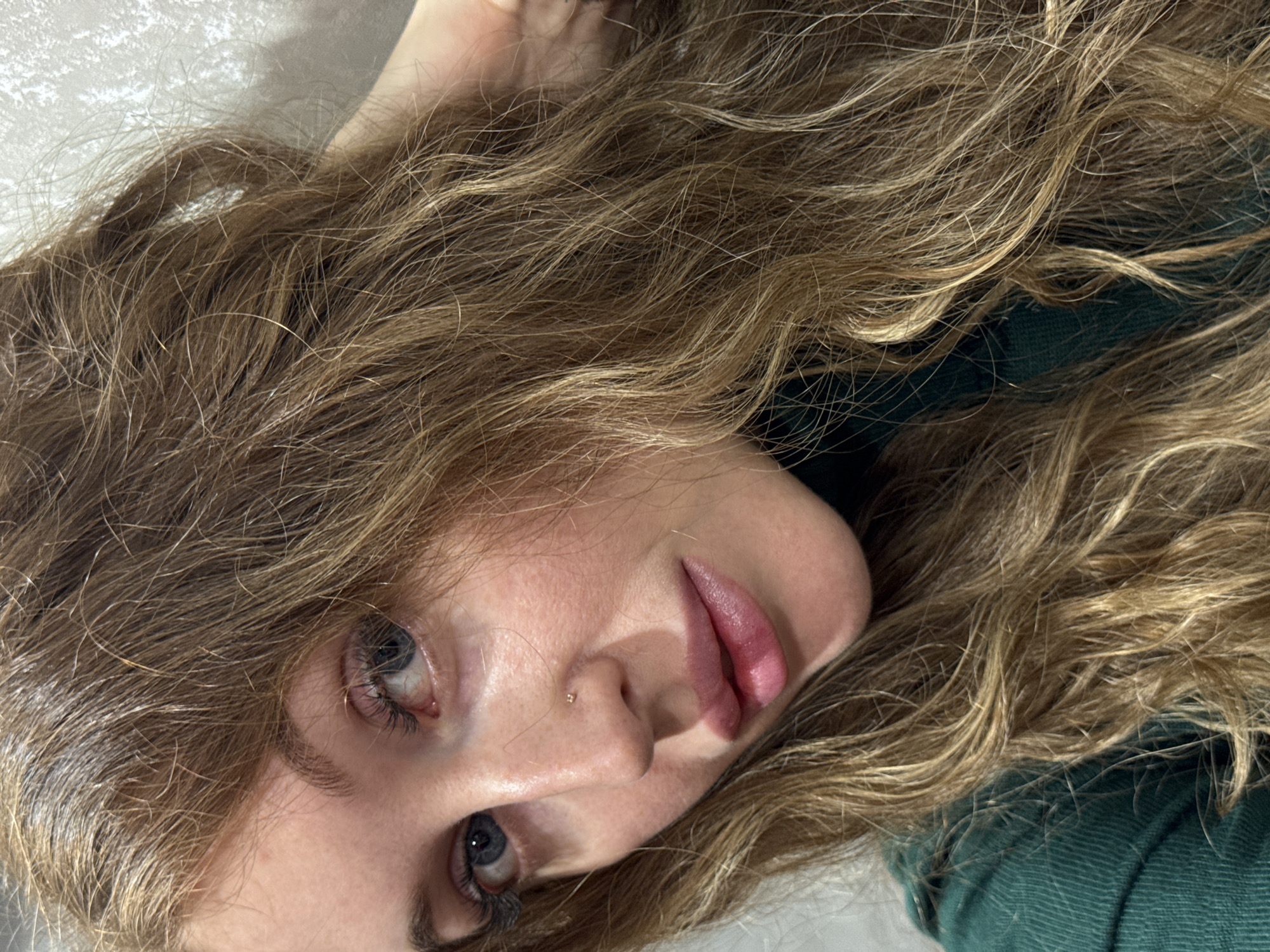 A close-up, tilted, angled shot depicts a woman with long, wavy blonde hair. Her face is prominent, revealing clear skin, full lips, and light blue eyes. A small gold nose stud is visible on her left nostril. She is wearing what appears to be a dark green top. The background is a soft, out-of-focus white. The location of the image in the city of Chaikovsky, Russia, is not discernible from the visual information provided.