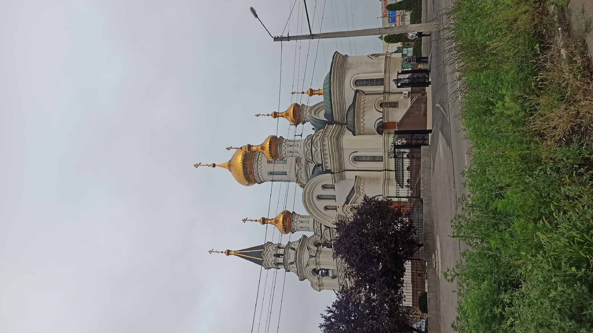 A wide shot captures an Orthodox church with multiple golden domes under a gray, overcast sky. The church is white with architectural details and crosses atop its spires. To the left of the church, a dark purple-leaved tree stands behind a black metal fence. In front of the church, a dark asphalt road curves, bordered by a stretch of green grass and weeds. A tall, gray lamppost stands to the right of the church, with power lines crossing the frame. A few indistinct figures are visible near the entrance of the church, and in the background, there are buildings and trees.
