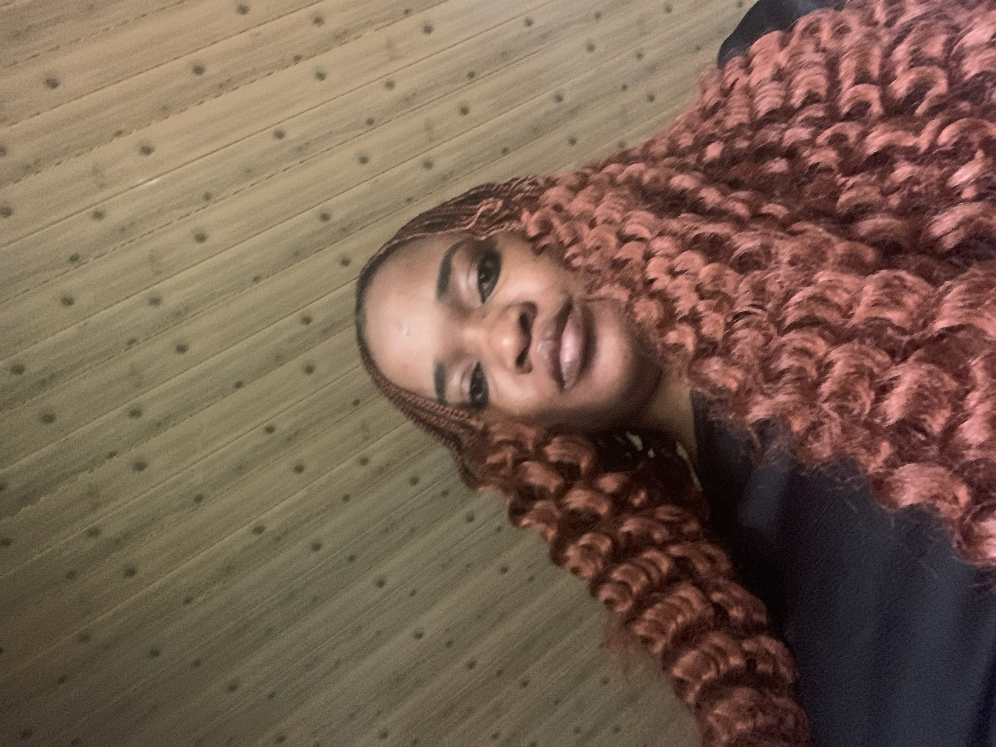 A young woman with dark skin and brown curly hair lies on a wooden floor. She is wearing a dark-colored shirt. The location of the scene is unknown.