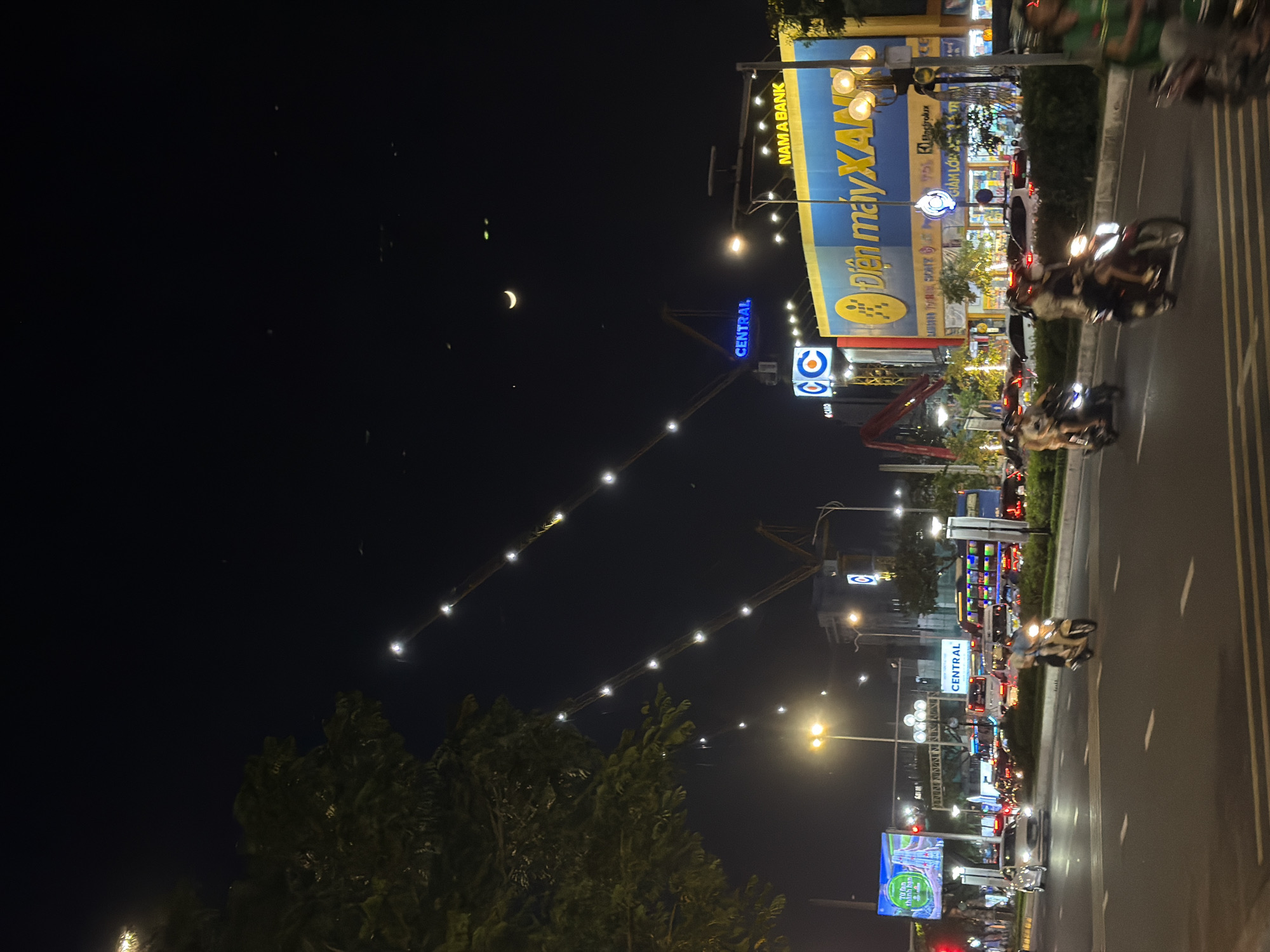 The image depicts a nighttime street scene in Đà Nẵng, Vietnam. A busy street is lined with buildings adorned with brightly lit signs, including one for "Điện máy XANH" and "NAMA BANK." Traffic consists mainly of scooters and motorcycles moving along the road, their headlights blurring slightly due to motion. Overhead, construction cranes are illuminated with strings of lights. In the night sky, a crescent moon is visible. The image also captures details of roadside advertising and the general urban landscape.
