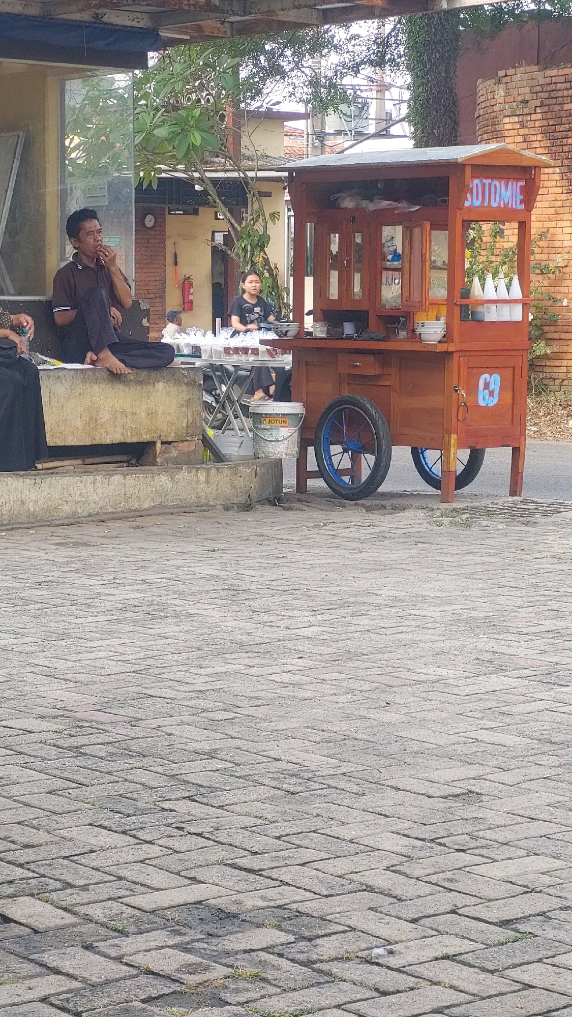 In Bojong Gede, Indonesia, a street food cart vendor is preparing food from their wooden cart. The cart has the word "SOTOMIE" in white lettering on its roof. A young woman is behind the cart, and to the left, a man sits with his legs crossed, smoking. Several customers are visible, and the cart is situated on a brick-paved surface adjacent to a road. A white bucket with the brand name "JOTUN" is visible near the cart. The scene is outdoors, with trees and buildings in the background.