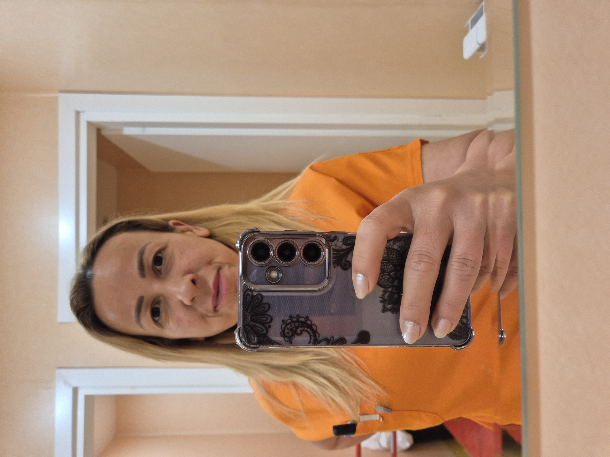 In an indoor setting, a person is taking a mirror selfie. The person, with blonde hair and fair skin, is wearing an orange shirt. They are holding a smartphone with a decorative case in their right hand, with their left hand resting on the phone. The background shows a peach-colored wall and white door frames. There are reflections in the mirror, including what appears to be part of a red chair and other objects. The overall impression is that of a casual, personal moment captured at home or in a similar private space.