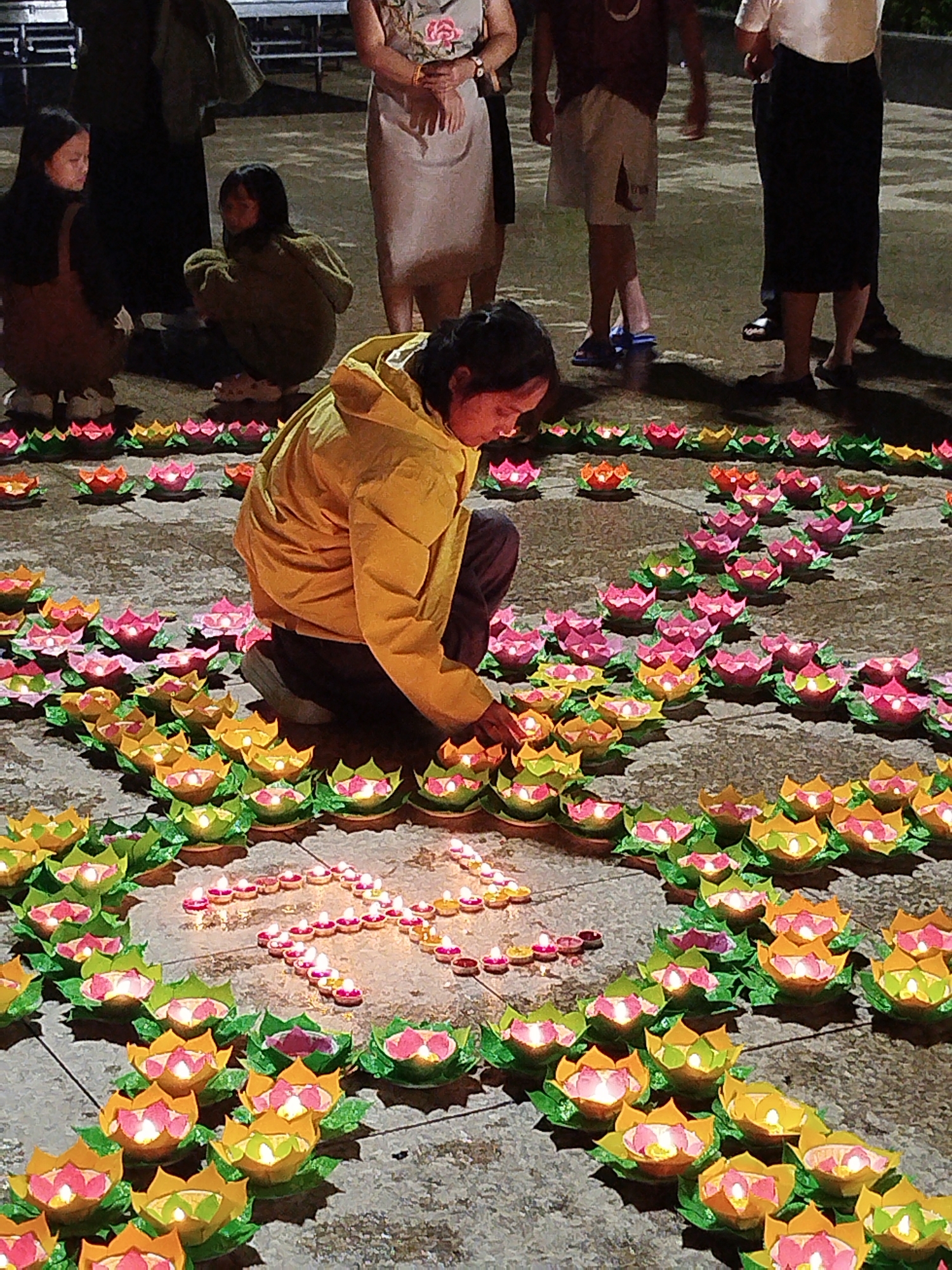 The image depicts a woman kneeling among rows of colorful lotus-shaped candle holders on a paved surface, likely a square or plaza. The woman wears a bright yellow jacket and appears to be lighting or adjusting a candle within the arrangement. The candle holders are arranged in rows and also form a cross shape made of individual tea light candles. Other people are visible in the background, some standing and some sitting. The scene appears to be set at dusk or night, given the darkness and the illumination from the candles. The location is the city of Da Lat, Vietnam, known for its Buddhist temples and cultural festivals that often involve the lighting of candles and paper lanterns.