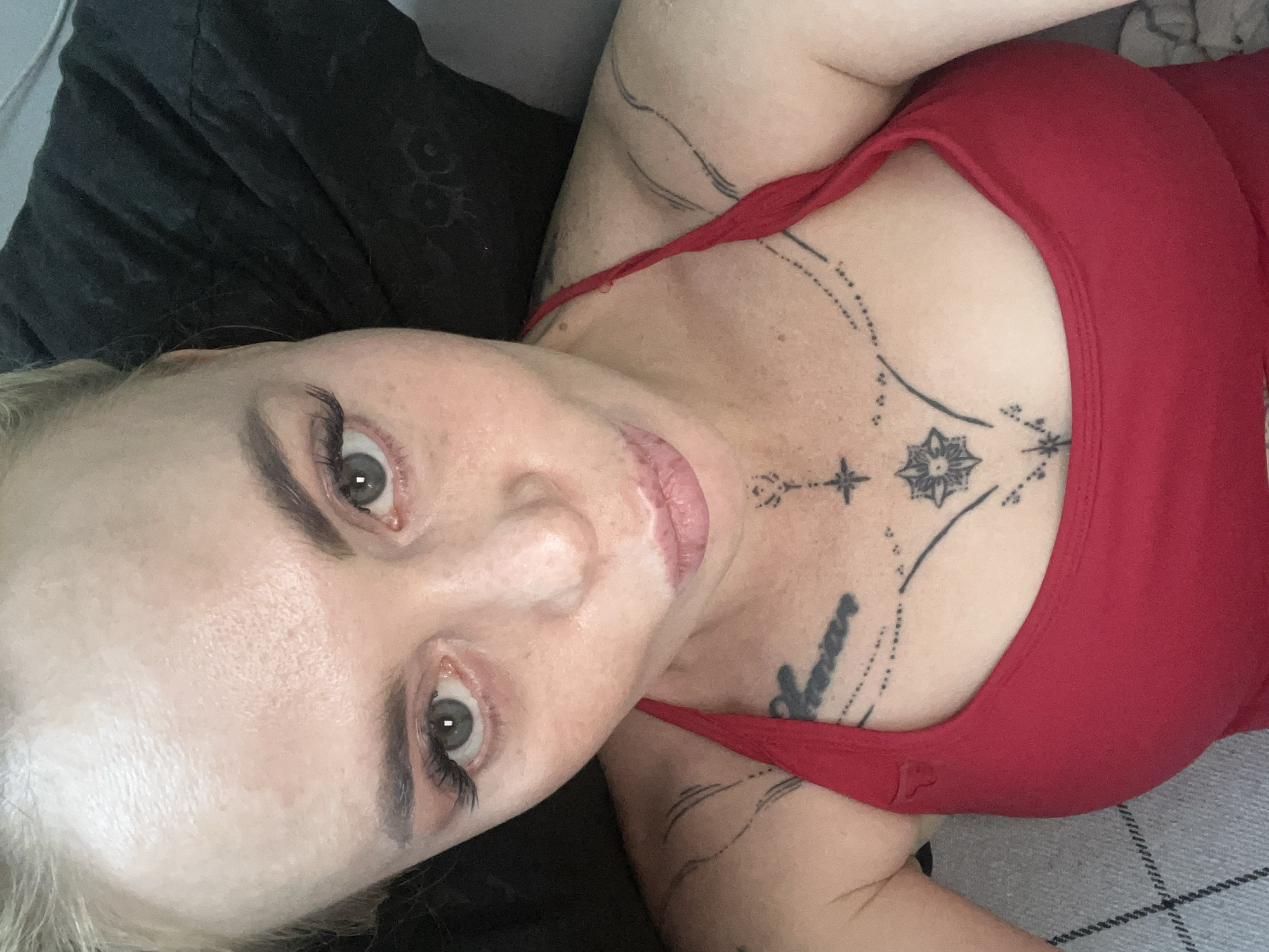 A close-up, high-angle selfie features a woman with fair skin, blonde hair pulled back, and light green eyes. She is wearing a red top and has visible tattoos on her chest and upper arms. Her lips are slightly pursed, and she is wearing makeup, including false eyelashes. The background consists of dark, textured fabric, possibly a pillow or blanket, and a hint of a gray surface. The lighting appears to be natural. The image is cropped to focus on the woman's face, upper torso, and shoulders. The scene is indoors, with no discernible urban environment or specific city landmarks.