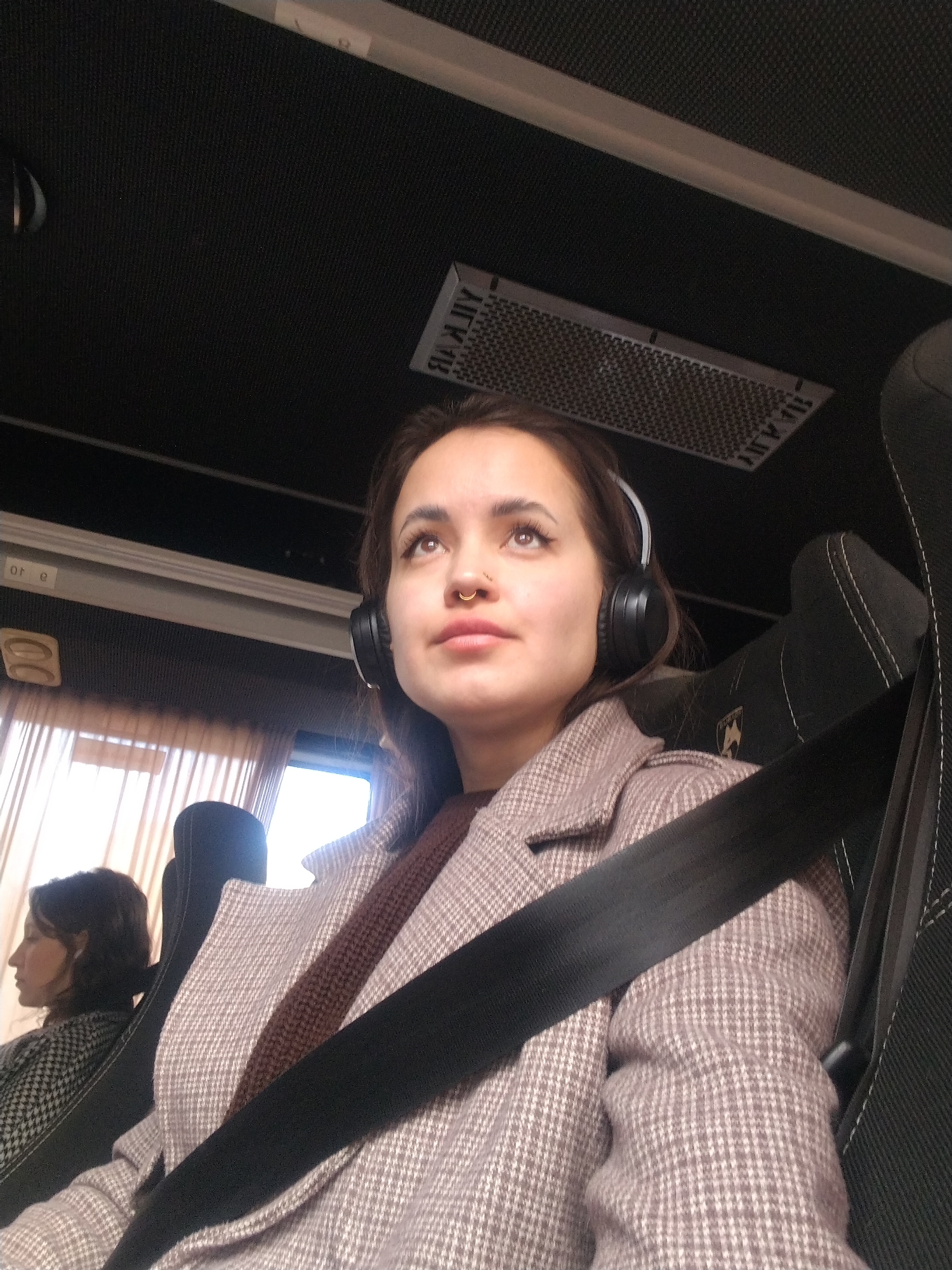 The image captures the interior of a passenger vehicle, identified as being within the city of Chaikovsky, Russia. The primary subject is a young adult female occupying the foreground, seated with a black seatbelt secured diagonally across her chest and lap. She has dark hair, a gold-colored septum ring, and is wearing black over-ear headphones. Her attire includes a light brown or beige houndstooth-patterned overcoat over a dark brown top. She gazes upwards and to her right with a neutral expression. To her left, partially visible, is another female passenger facing left, away from the camera, wearing a dark patterned garment. The vehicle's interior features dark, possibly black or dark gray, upholstered seats with visible white stitching on the headrest adjacent to the main subject. This headrest also displays a light-colored emblem. The ceiling is dark and textured, bordered by a light-colored trim. Overhead, an air vent with inverted Cyrillic text (e.g., "А1К1В", "ДАЙ Ж") is discernible. Windows on the left side of the vehicle are covered by light-colored curtains, with daylight visible beyond. The scene indicates travel during daytime.