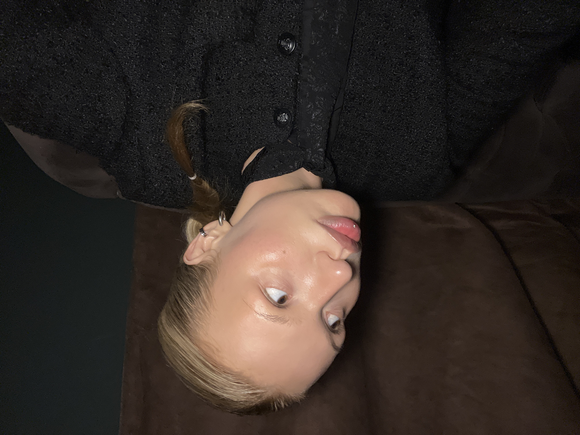A person with fair skin, light blonde hair, and brown eyes is looking upwards. The person is wearing a black textured top with buttons. Their hair is partially pulled back in a small ponytail. They appear to be lying on a brown surface. The image is rotated. The location in Chaikovsky, Russia is unknown from the image.