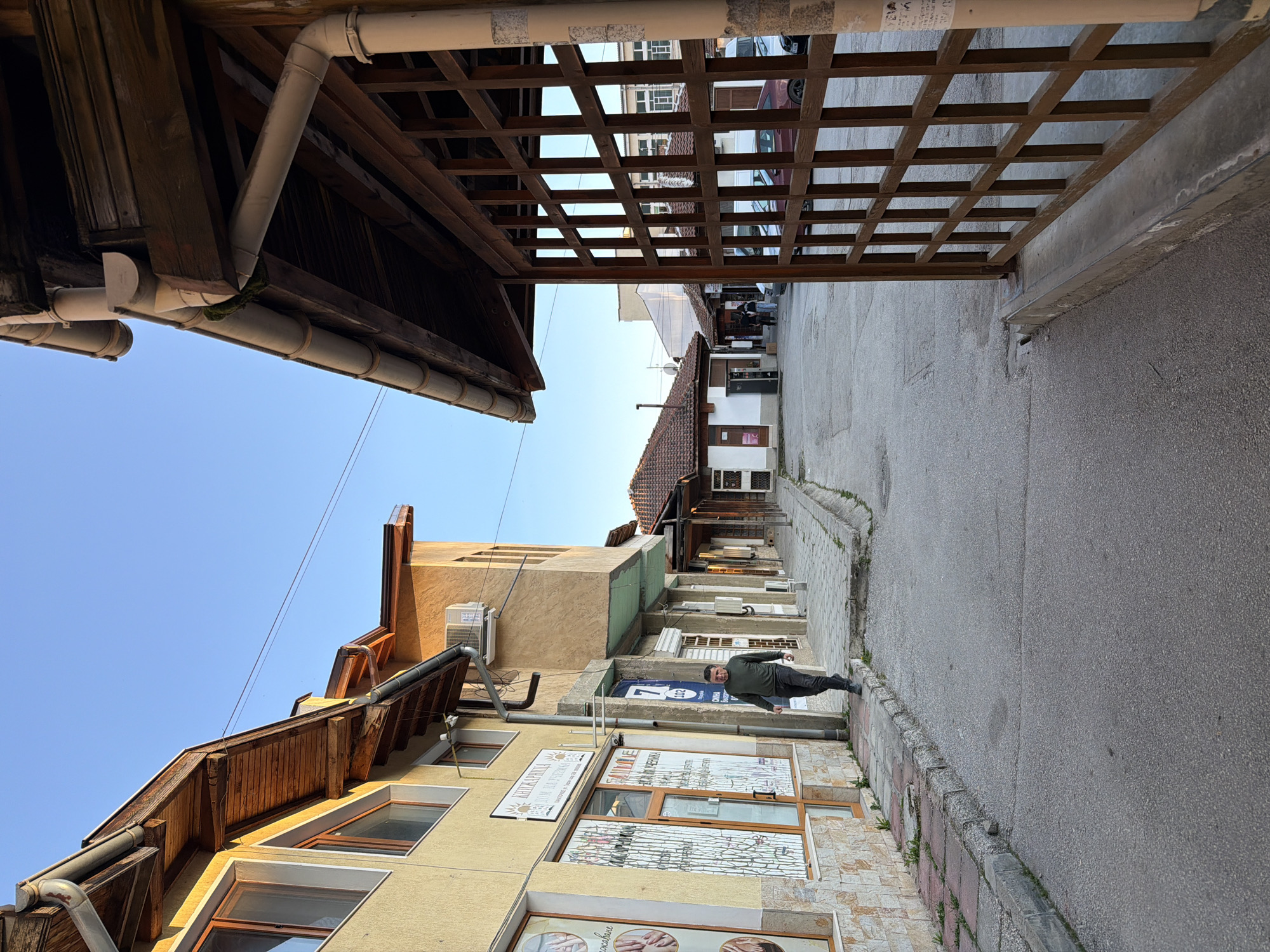 A street scene shows a person walking on a sidewalk alongside a narrow street with buildings on one side and a building with a wooden lattice roof on the other. The person is wearing dark pants, a long-sleeved dark shirt, and shoes. The buildings vary in height and color, with some having tile roofs. There are commercial signs and windows on the buildings. The street is gray asphalt with a curb. The sky is visible above the buildings and is a pale blue. The location is Panagyurishte, Bulgaria.