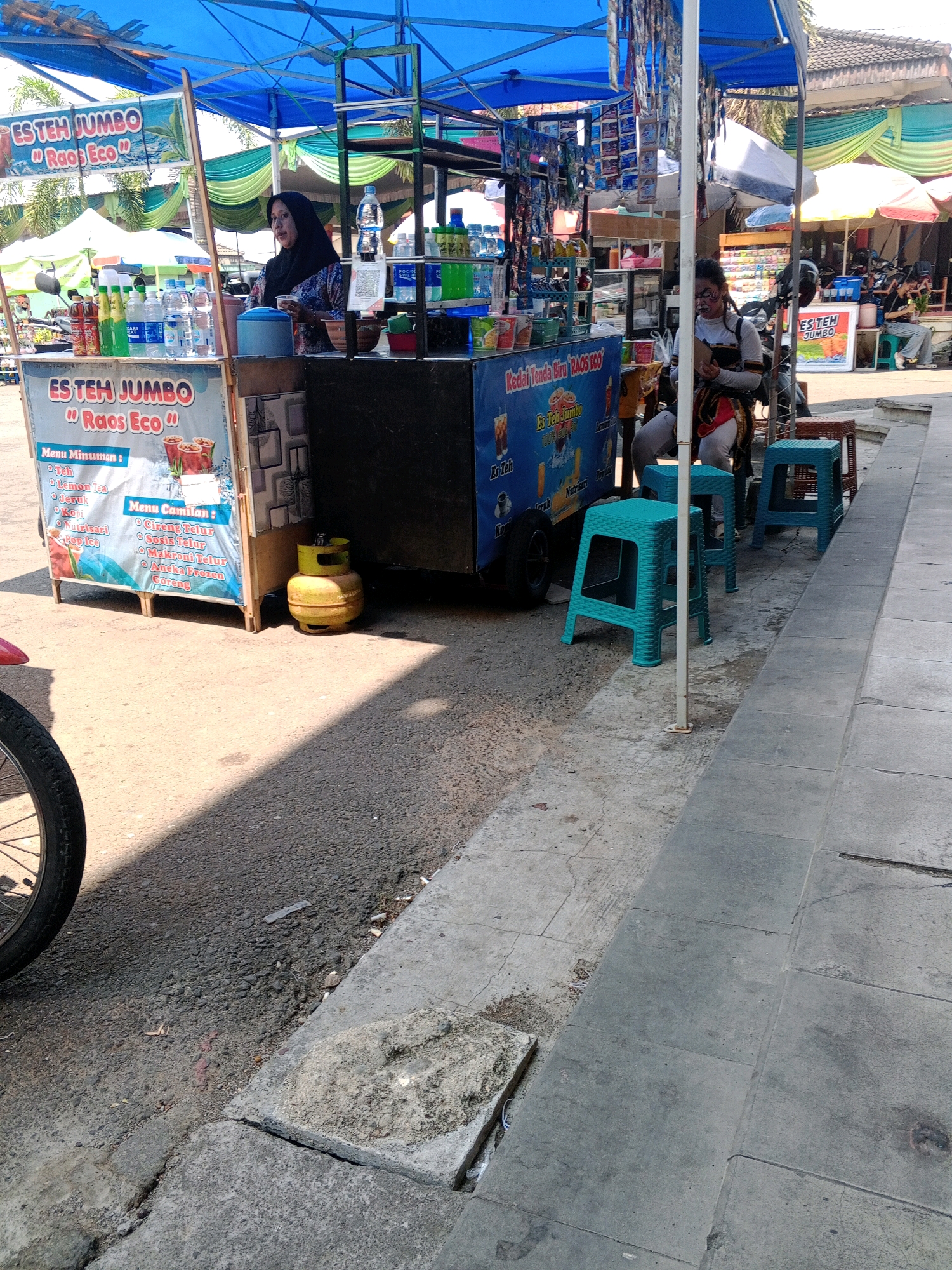 The image depicts an outdoor street scene in Wuryorejo, Indonesia, featuring a mobile food and beverage stall. A woman wearing a hijab and a patterned shirt is standing behind the stall, appearing to serve a customer. In front of the stall, several plastic stools are arranged, with one teal stool prominently in the foreground. The stall itself is blue and features signage with Indonesian text, including "ES TEH JUMBO" and menu items. A yellow propane tank is visible next to the stall. In the background, other stalls and people can be observed, suggesting a market or busy street. The ground is paved with asphalt and concrete.