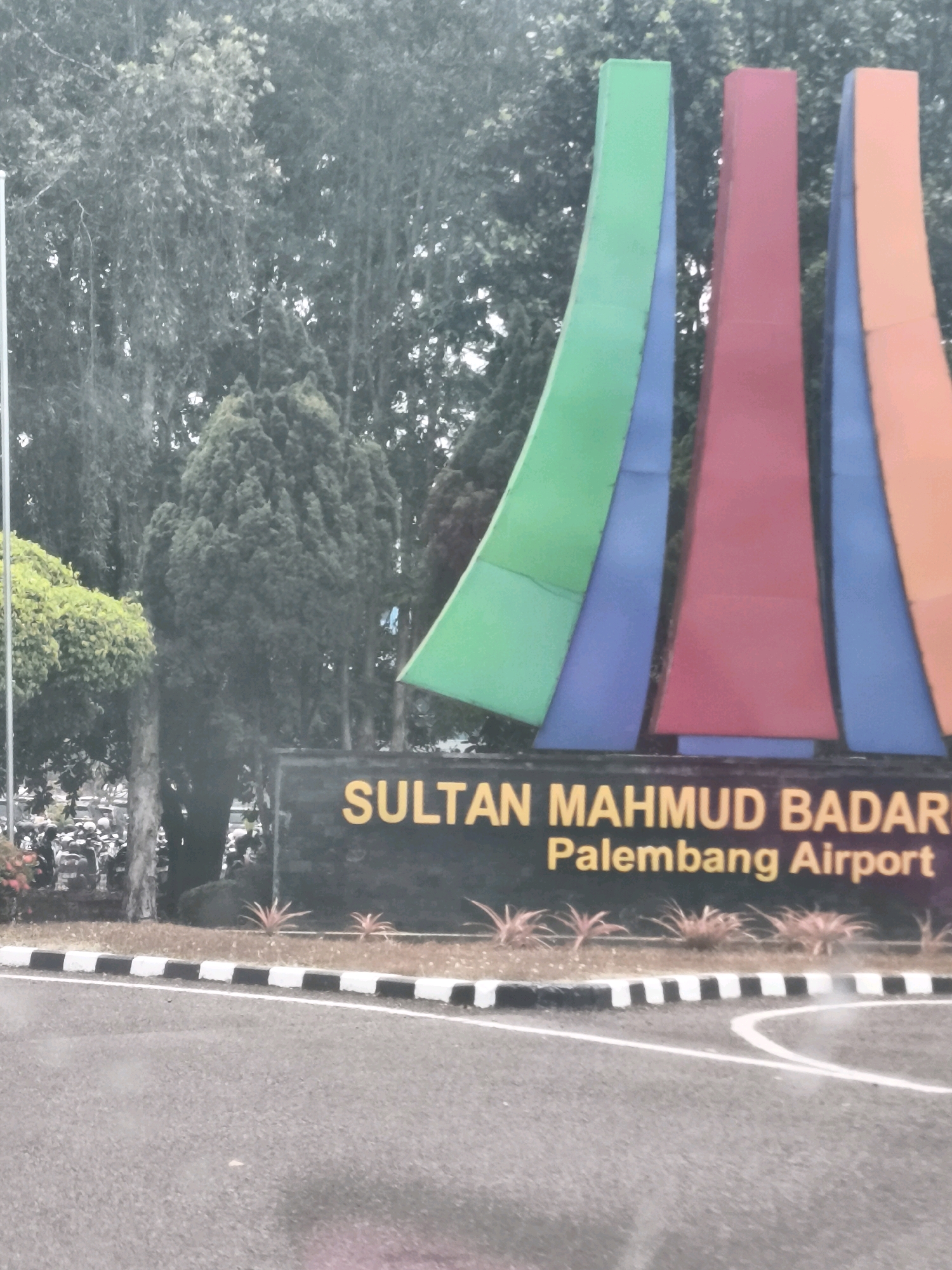 The image depicts the entrance to Sultan Mahmud Badaruddin II International Airport in Palembang, Indonesia. In the foreground, a paved road curves, bordered by a black and white striped curb. Behind the curb, a strip of dry, sparse vegetation with pinkish-red leaves is visible. Dominating the background is a large, dark sign with yellow lettering that reads "SULTAN MAHMUD BADARUDDIN II" on the top line and "PALEMBANG AIRPORT" on the bottom line. To the left of the sign, a dense cluster of trees and foliage forms a green backdrop. Directly behind the sign, a series of tall, colorful, abstract structures rise vertically, with colors including green, blue, red, and orange. To the far left, a white pole with a white flag is partially visible. Further left, beyond the pole and a bit of foliage, a group of motorcycles and people are visible in what appears to be a parking area. The overall scene suggests an airport entrance under overcast skies.