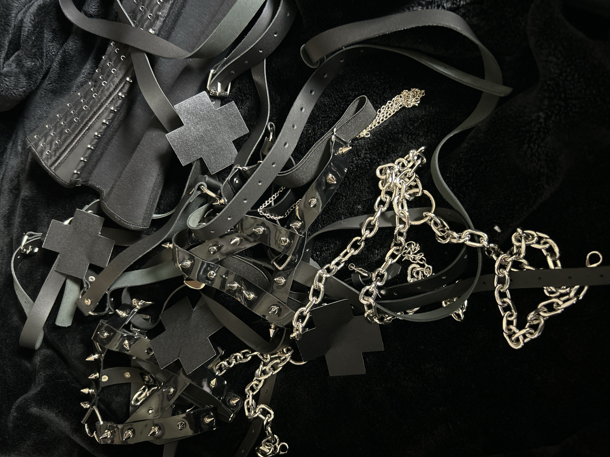 The image depicts a collection of black gothic fashion accessories arranged on a black, plush surface. The items include a studded corset, harnesses with metal crosses, studded belts, and silver chains. The setting appears to be a studio or indoor space due to the controlled lighting and background. The location, Cape Coral, United States of America, is not evident in the image.