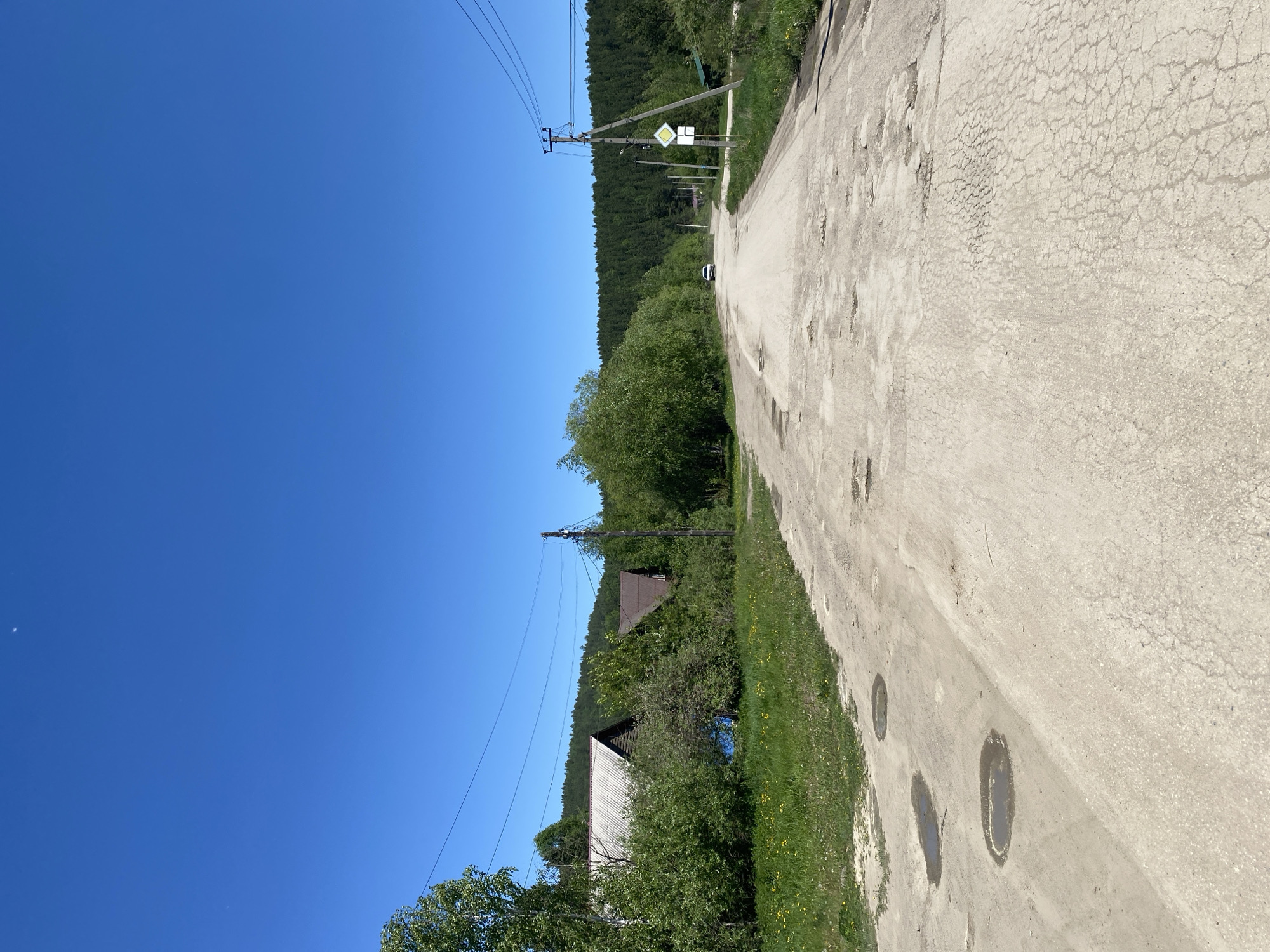 The image depicts a dirt road in the village of Bolshoy Lug, Russia, on a clear, sunny day. The road is unpaved and shows signs of disrepair, with numerous potholes and patches of cracked asphalt. To the left of the road, a grassy embankment is visible, dotted with yellow wildflowers and a few puddles. Beyond the embankment, several houses are partially obscured by lush green trees and bushes. The houses appear to be of traditional wooden construction, with one having a white corrugated metal roof and another a darker, possibly wooden, roof. Power lines run overhead, supported by a wooden pole on the right side of the road. A white car is seen driving away in the distance, kicking up a faint dust trail. The road curves gently to the right, leading towards a dense forest that forms the backdrop of the scene. The sky is a vibrant, cloudless blue.