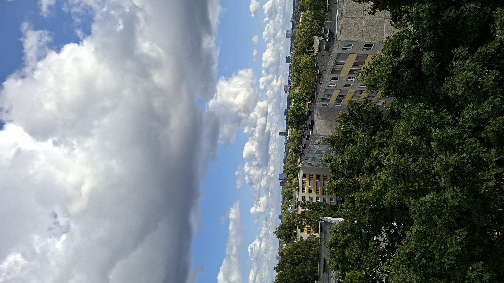 The image shows a cityscape with multiple apartment buildings and dense green trees. The sky is partly cloudy with large, dramatic clouds dominating the upper portion of the frame, interspersed with patches of blue sky. In the foreground, lush green trees obscure the lower parts of some buildings, suggesting a natural element integrated into the urban landscape.
Behind the trees, several apartment buildings are visible. Some are mid-rise with light yellow balconies or accents, while others are grey concrete structures. The roofs of these buildings appear to be flat, with visible ventilation units or other rooftop structures. Further in the distance, more buildings and trees form a layered cityscape, with some taller structures and a faint industrial element, possibly a smokestack, visible on the horizon. The overall impression is that of a residential area within a city, with a significant amount of greenery.
Based on the architectural style of the buildings, particularly the apartment blocks, and the urban planning with green spaces, this scene is characteristic of Soviet-era housing estates found in many Eastern European cities. The specific architecture of the apartment buildings, with their repetitive balconies and concrete facades, strongly resembles those found in areas like Lasnamäe in Tallinn, Estonia. The presence of distant skyscrapers also aligns with the urban development patterns of modern Tallinn. The image captures a typical view of a residential district in Tallinn on a cloudy day, with a mixture of aging Soviet-era architecture and the newer urban development on the horizon.