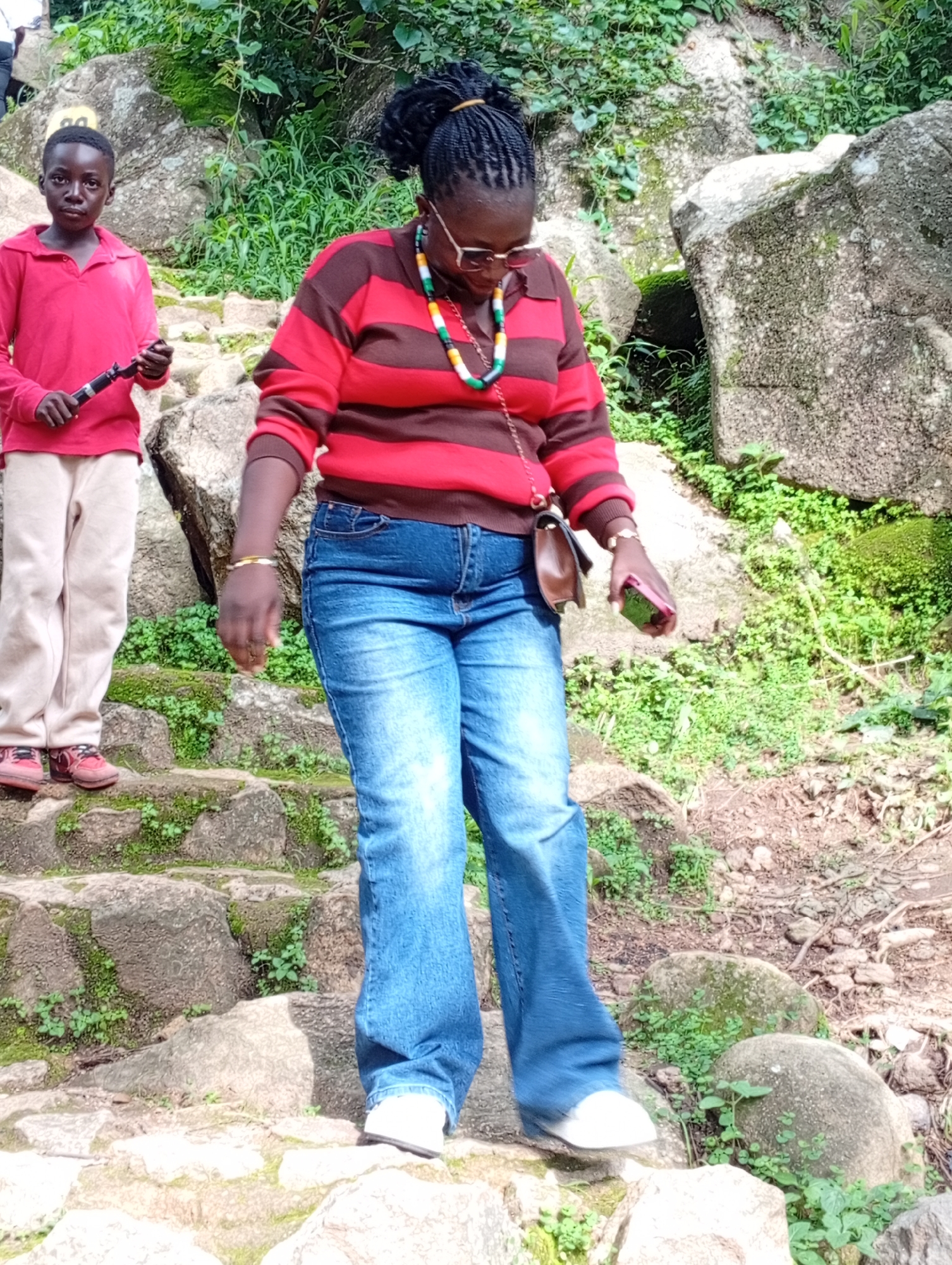 Two individuals are walking down a stone staircase in an outdoor, natural environment. On the left, a young Black person in a red collared shirt and light-colored pants holds a black object, possibly a clarinet or a similar instrument, and wears red shoes. To the right, a Black adult, also on the staircase, is dressed in a red and brown striped long-sleeved shirt, blue jeans, and white shoes. They are wearing glasses, a colorful beaded necklace, a wrist watch, and a brown purse is slung over their right shoulder. The adult is holding a pink mobile phone in their right hand. The staircase is made of rough, irregular stones, with moss and small green plants growing in the crevices. Large rocks and dense green foliage are visible in the background, suggesting a natural, possibly wooded or mountainous, setting. The lighting indicates it is daytime.