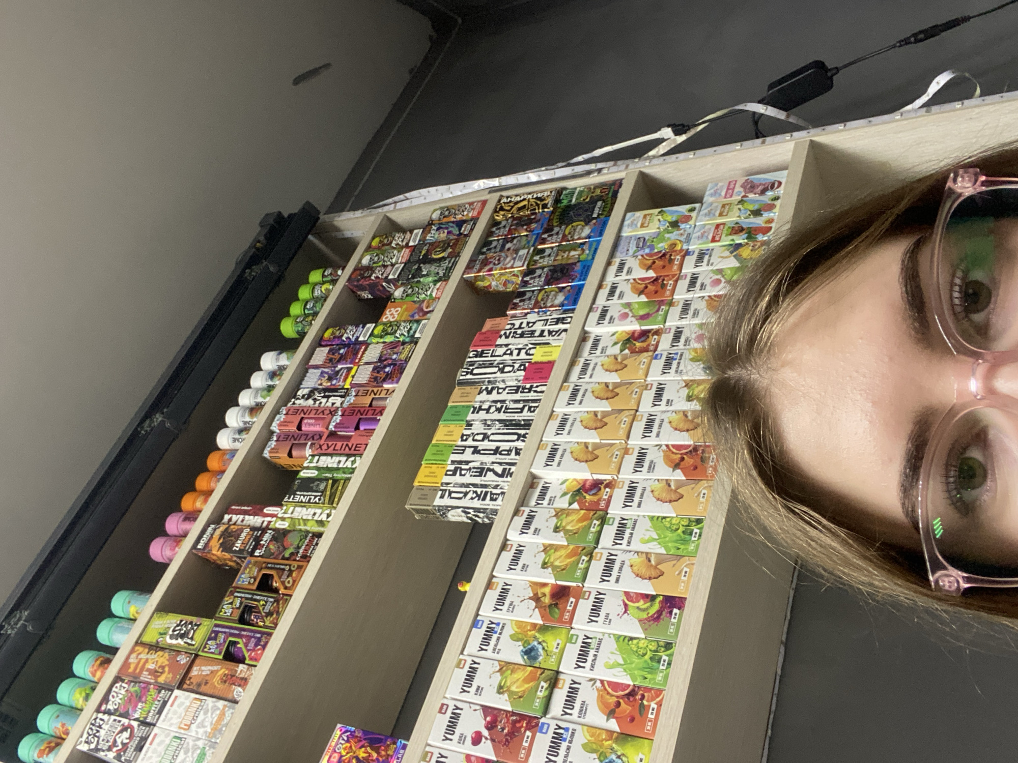 A close-up, slightly upward-angled view captures the upper portion of a person's face positioned in front of multi-tiered retail shelving in Дьяково, Russia. The individual, identified by light brown hair, green-hazel eyes, and pink-rimmed glasses, occupies the lower third of the frame, gazing directly at the merchandise. The background is dominated by a light-colored wooden shelving unit displaying numerous products, predominantly packaged e-liquids or disposable vape devices. The top shelf holds various small, colorful plastic bottles (green, pink, orange, white) and boxes bearing brand names such as 'GORBANI CRITICAL,' 'Waffle Cup,' 'RONIN,' 'ZAKURO,' and 'XYLINET,' featuring graphic designs including skulls. The middle and lower visible shelves are densely packed with 'YUMMY' branded boxes, each illustrated with different fruit flavors like cola, kiwi, orange apple, grape, pear, sour pineapple, and pina colada. A white LED light strip runs vertically along the right side of the shelving unit, with a connected black power adapter and cables. Above the top shelf, a black track or vent structure is visible, leading to a light gray ceiling in the upper right corner. The wall behind the shelves is a dark gray. The scene suggests an interior retail environment, likely a vape shop.