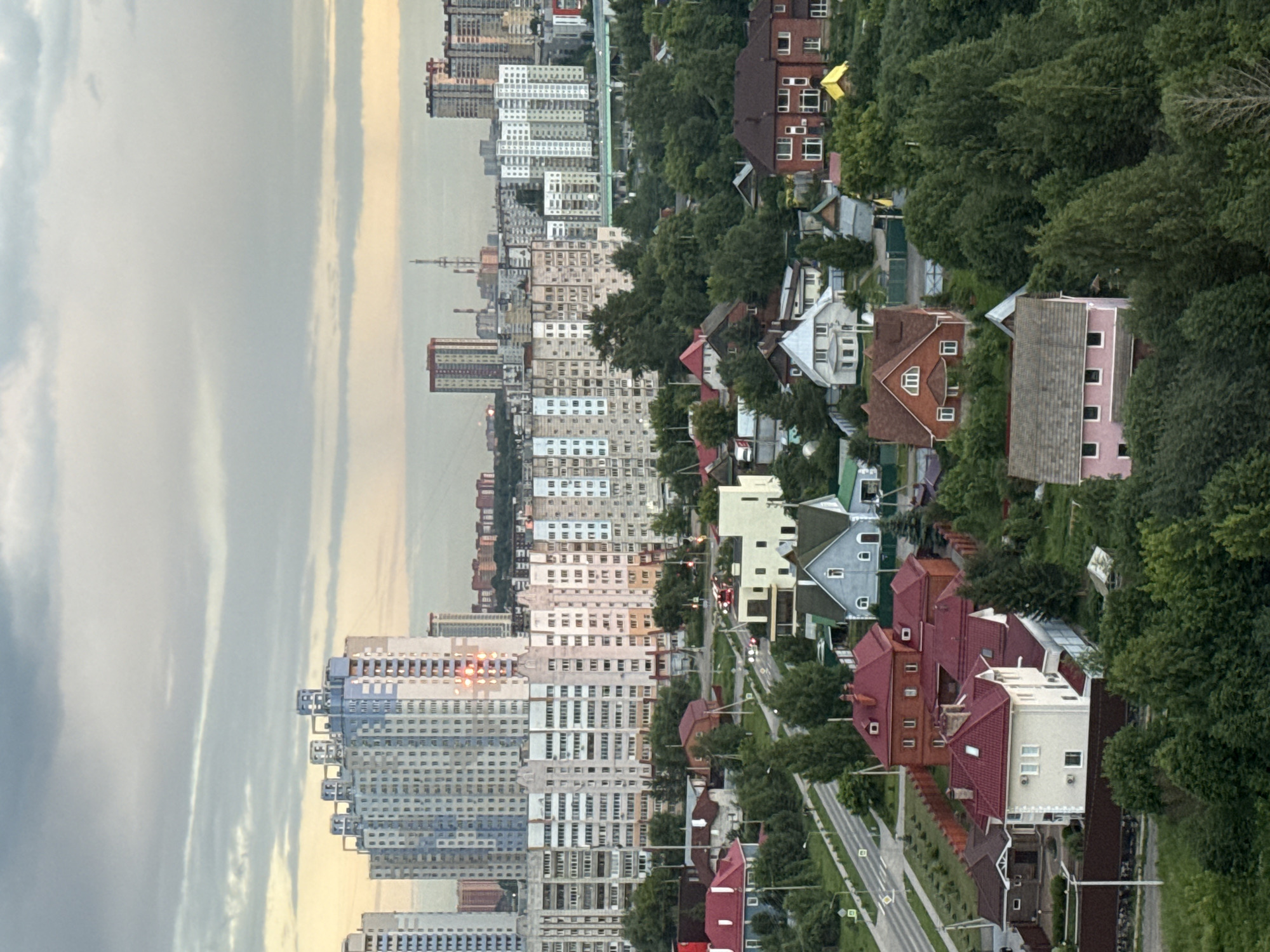 The image shows an aerial view of a suburban neighborhood in Perm, Russia, characterized by a mix of high-rise apartment buildings and individual houses. In the foreground, several detached houses with pitched roofs are visible, some featuring brick facades and others painted in lighter colors. These houses are surrounded by green trees and vegetation. A paved road runs through the scene, with a few cars present. In the background, a dense cluster of multi-story apartment buildings dominates the skyline, with some featuring light blue and white exteriors. The sky is overcast with soft, diffused light, suggesting it might be early morning or late afternoon. The overall impression is of a developing urban area with a blend of residential types.