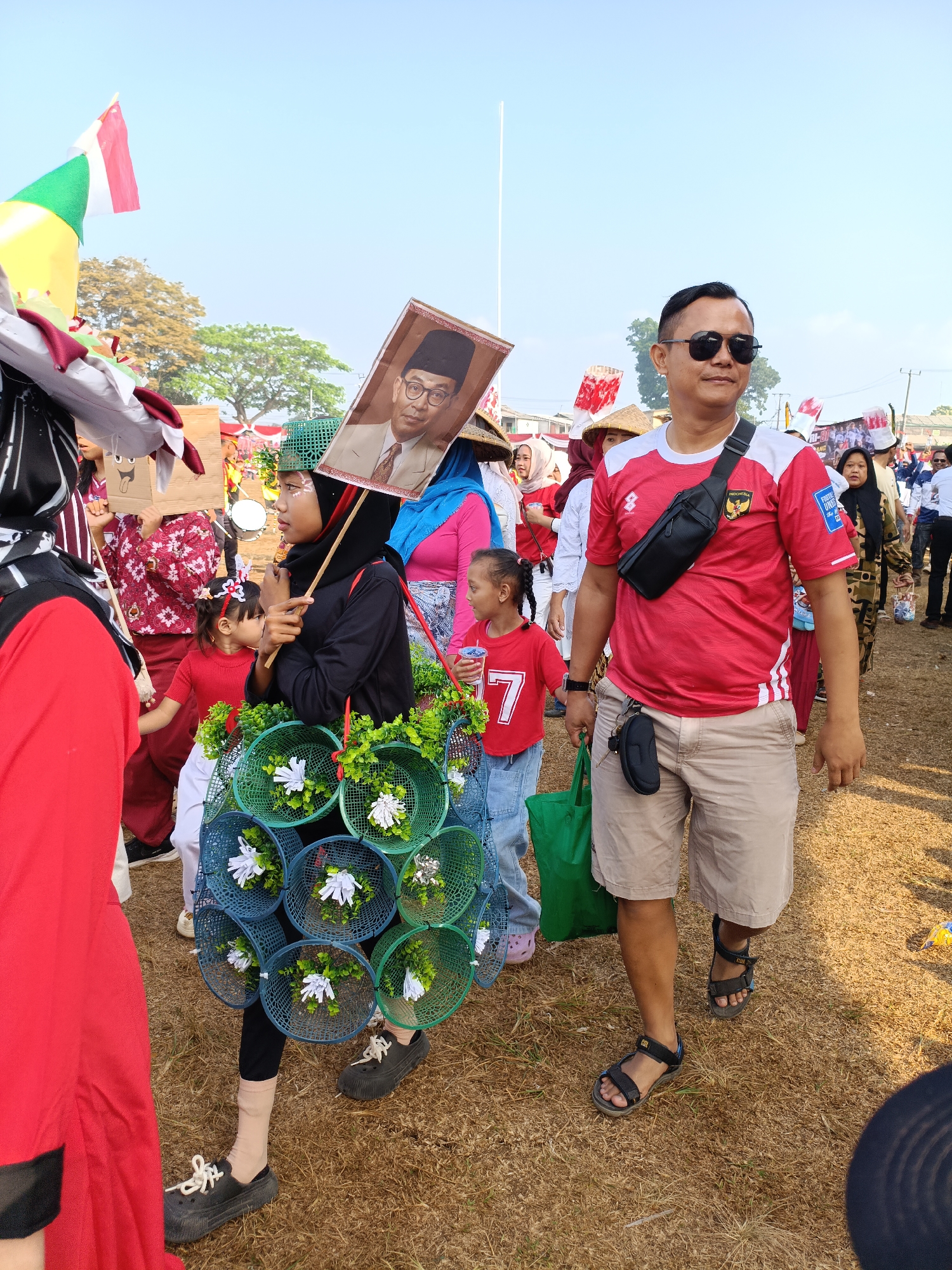 In Cibodas, Indonesia, a group of people are participating in an outdoor parade. A man in a red and white shirt and beige shorts walks towards the camera, wearing sunglasses and a black fanny pack. To his left, a young girl in a red shirt with a white number "7" on it walks, holding a drink. Behind her, a woman wearing a black hijab and a black long-sleeved shirt is carrying a large circular ornament adorned with greenery and white flowers. She is also holding a portrait of a man in glasses and a suit. Other individuals, some in traditional Indonesian attire, are visible in the background, along with flags and tents. The ground is covered in dry grass, suggesting a warm, outdoor environment.