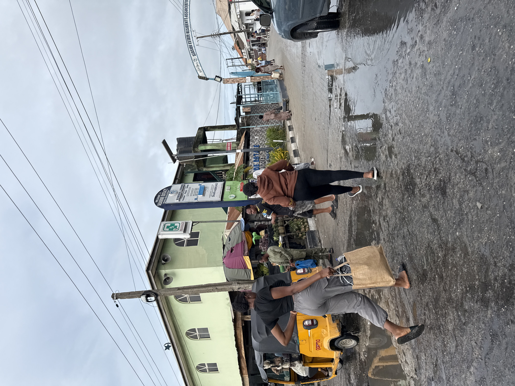The image captures a street scene in Ado, Nigeria, likely after a rain. The wet pavement reflects the surroundings. Two individuals walk along the road; one carries a brown tote bag, the other wears a brown top and black leggings. A yellow tricycle ("Keke Napep") is parked nearby. Fruit sellers with pineapples and bananas are visible along the side of the road. Buildings include a pharmacy and D'Cherub schools. A banner for "Seaside Estate Residents Association" is strung across the street. Overhead, utility wires crisscross the sky.