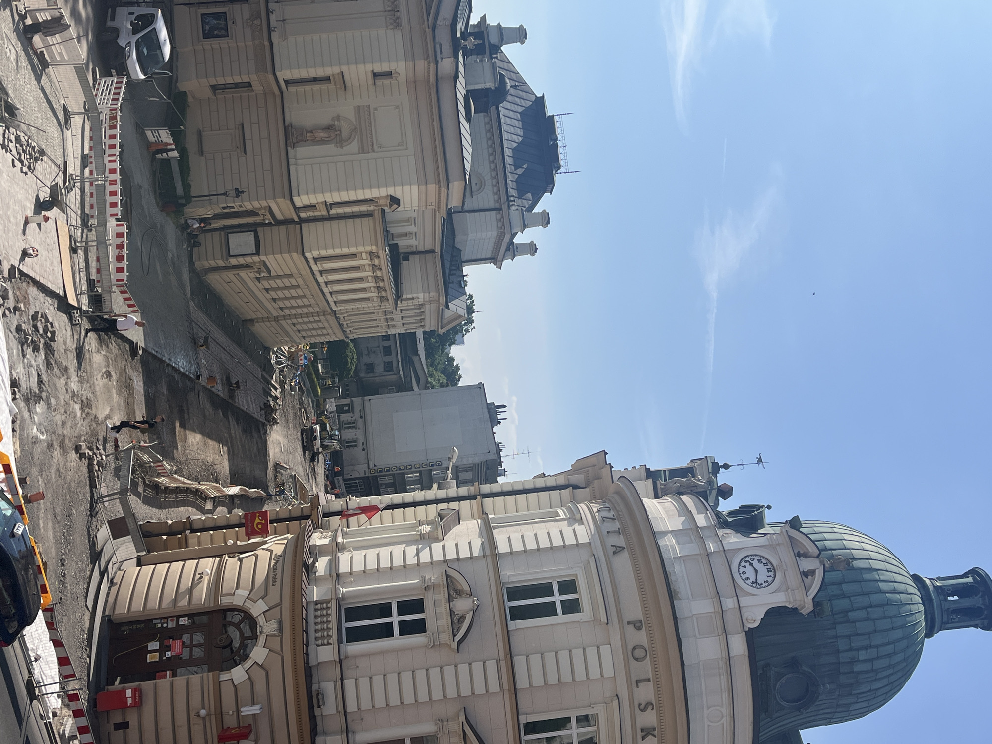The image shows a sunny day in an urban environment. In the foreground, there is construction activity on a street, with barriers, paving stones, and a white van. A person is standing near the barriers. Behind the construction, two large buildings are visible. The building on the right is light-colored with a prominent green dome and a clock. Text on this building reads "POCZTA POLSKA". The building on the left is also light-colored with a dark roof. In the background, other buildings and trees can be seen, as well as a clear blue sky with a small bird.