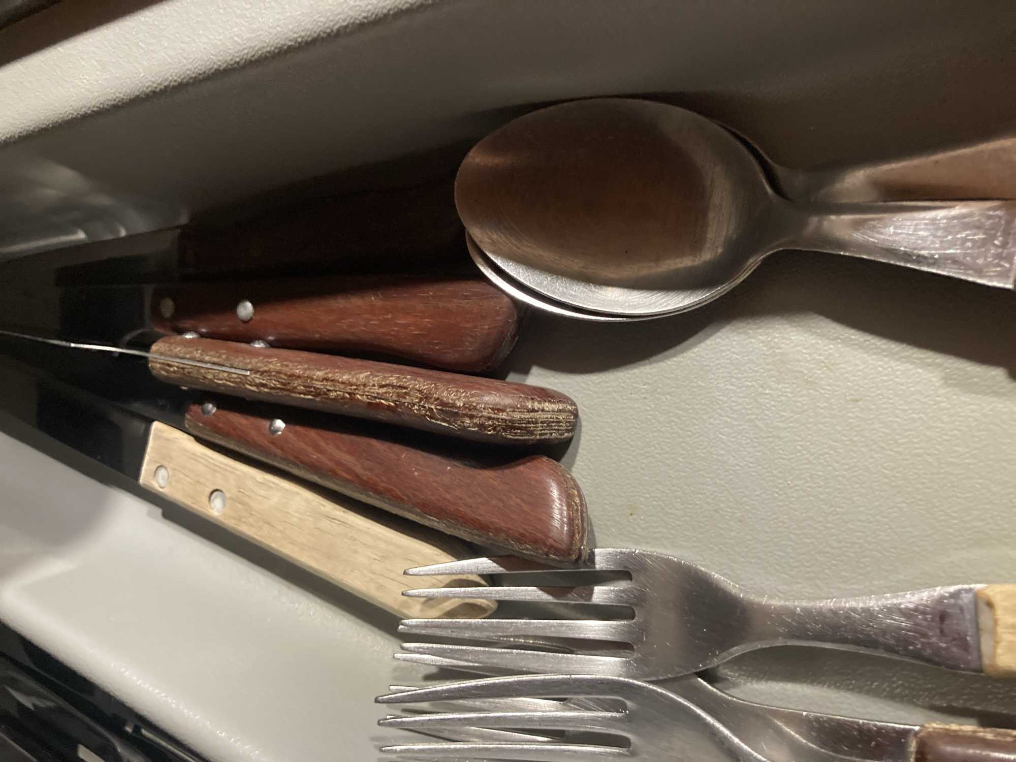 A collection of silverware, including knives, forks, and spoons, is neatly arranged in a gray utensil tray. The knives feature wooden handles in various shades of brown, with metal blades. The forks are made of polished metal. Two spoons are placed side-by-side, also made of polished metal. The scene is indoors, and the lighting suggests it could be within a kitchen or dining area. The exact location in Sabadell, Spain, is not discernible from the image.
