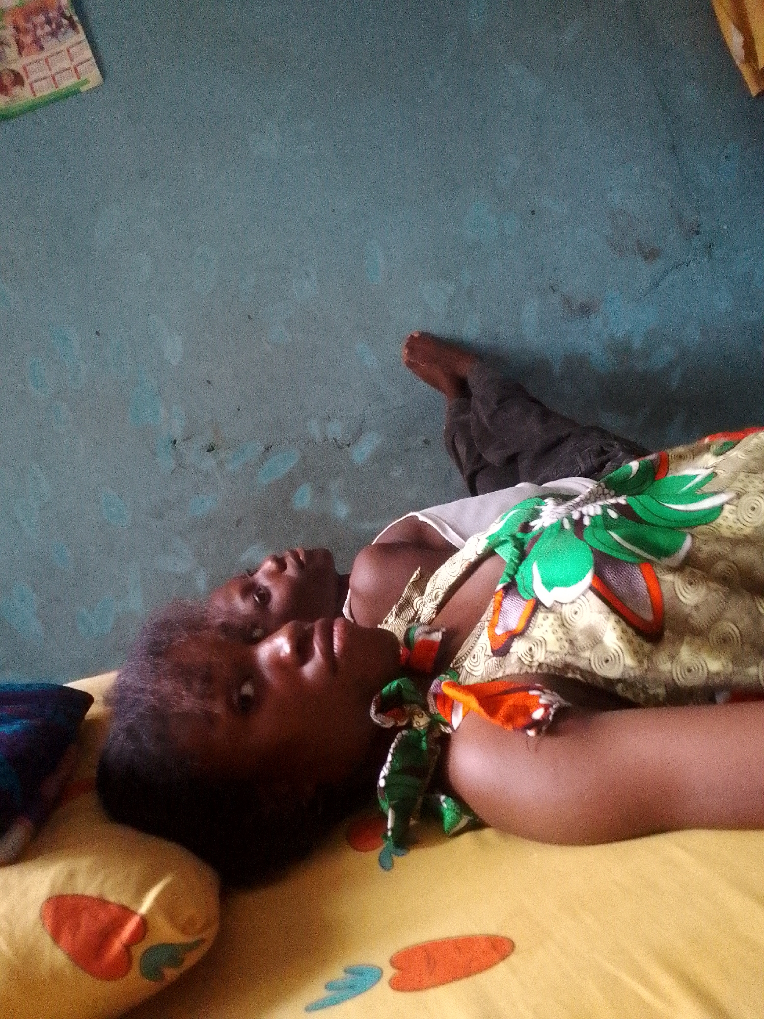 Two people lie on a yellow bedspread patterned with red and blue shapes. The bed is positioned against a blue wall. The person closest to the camera is a dark-skinned individual with short, dark hair, lying on a yellow pillow. Next to them, another person is lying, partially obscured. Their bare foot is visible near the top right of the frame. The visible person wears a white top and a patterned cloth with green, orange, and white floral-like designs. A small, green, doll-like object is also visible on the bed. The location is in the city of Unknown, Unknown.