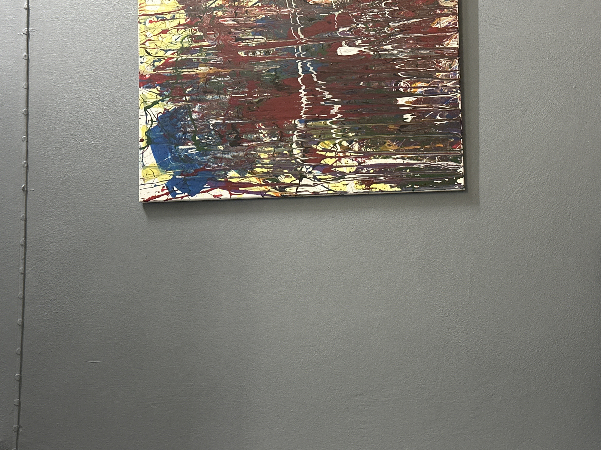 A close-up, eye-level shot captures a section of a gray painted wall in Chalong, Thailand, featuring an abstract drip painting. The painting, predominantly in hues of red, yellow, and blue, is positioned on the right side of the frame, with its left edge aligned approximately one-third of the way across the image. The artwork exhibits vertical streaks of paint, creating a sense of dripping motion. The wall itself has a slightly textured finish, and a horizontal line of small, evenly spaced rivets or nails runs across the top of the frame. The lighting appears to be indoor, casting subtle shadows. No people are visible in the image.