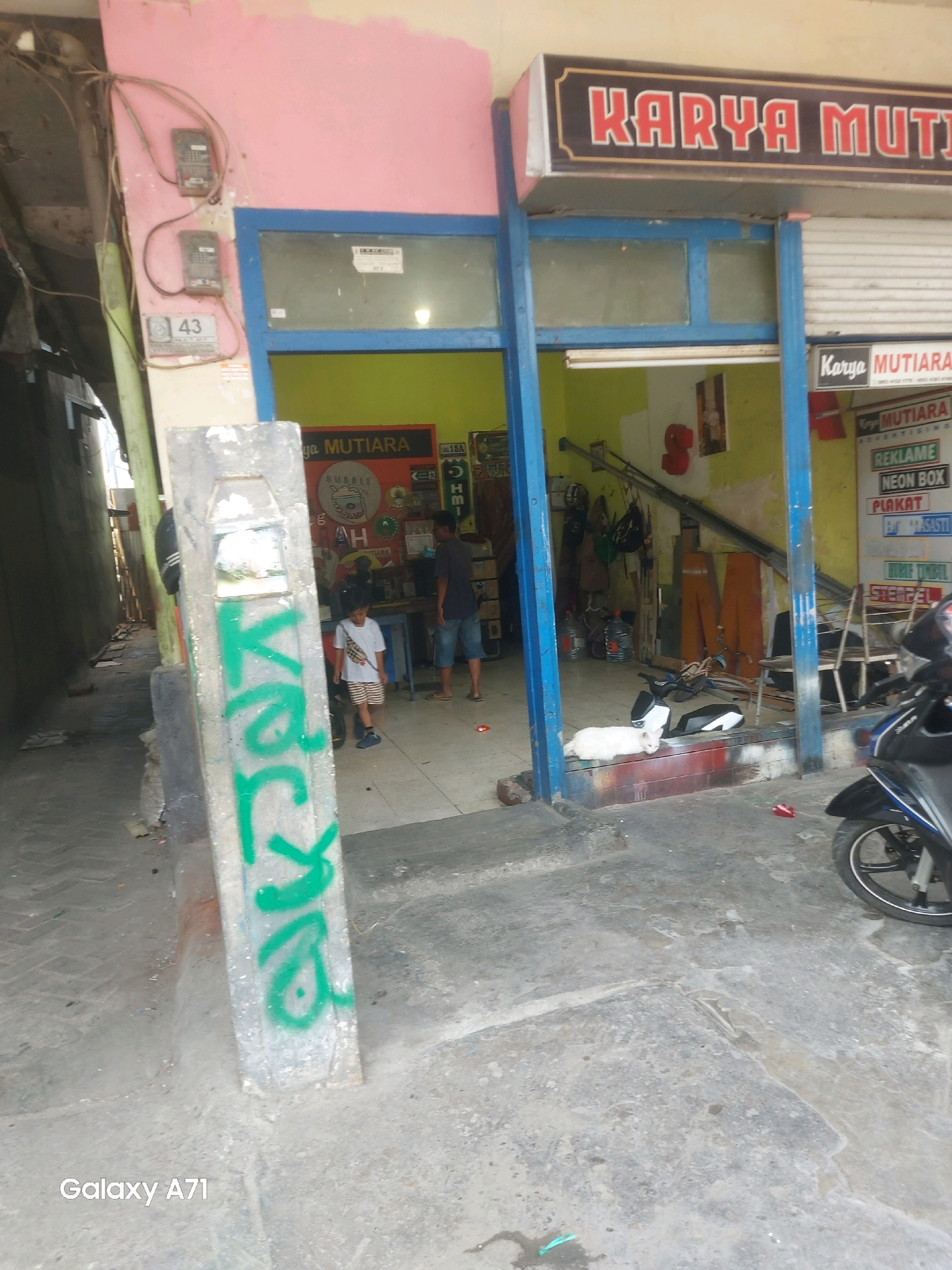 The image depicts the exterior of a commercial establishment identified as "KARYA MUTIARA" in Makassar, Indonesia. The storefront features a prominent sign with red lettering on a dark background, situated above blue-framed windows and a white roll-up door. Inside, the space appears to be an advertising or printing shop, with various materials and equipment visible, including what looks like large orange panels and metal shelving. A white cat is lying on the floor near the entrance. In the foreground, a concrete pillar is covered in green graffiti. Two people are present: a young boy in a white t-shirt and striped shorts standing near the entrance, and an adult male wearing a dark t-shirt and denim shorts standing further inside the shop. A motorcycle is parked to the right of the frame. Electrical meters are mounted on a pink wall to the left, along with a sign displaying the number "43". The overall environment suggests a bustling urban setting with a mix of commercial activity and street elements.