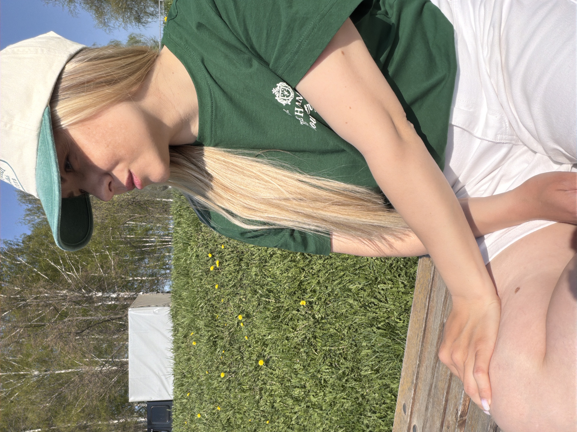 A young woman with blonde hair and fair skin is seated outdoors on a wooden bench in a grassy area dotted with yellow flowers. She wears a green t-shirt with a white logo on the left chest, white shorts, and a beige baseball cap with a green brim. Her hands rest on her lap. Behind her, a small white tent or structure stands amidst a backdrop of bare trees against a blue sky. The location is potentially a park or green space within the city of Votkinsk, Russia.