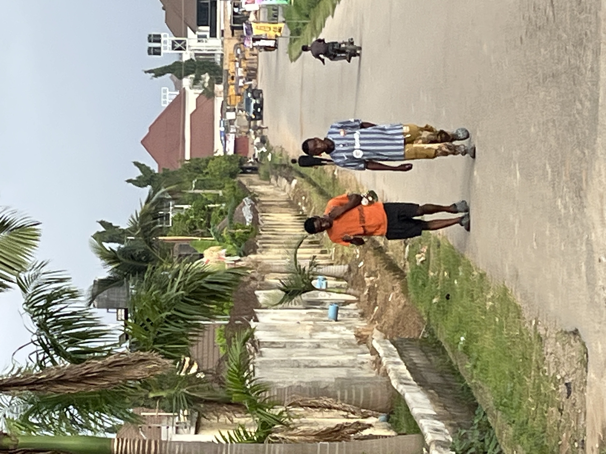 Two young men are walking down a paved road in Abuja, Nigeria. The man on the left is wearing an orange t-shirt and black shorts, while the man on the right is wearing a striped t-shirt and patterned pants. Both men are walking towards the camera. In the background, there are several buildings with red roofs, palm trees, and other vegetation. A motorcycle is being ridden by a person further down the road, and a dark SUV is also visible. The scene appears to be a residential area.