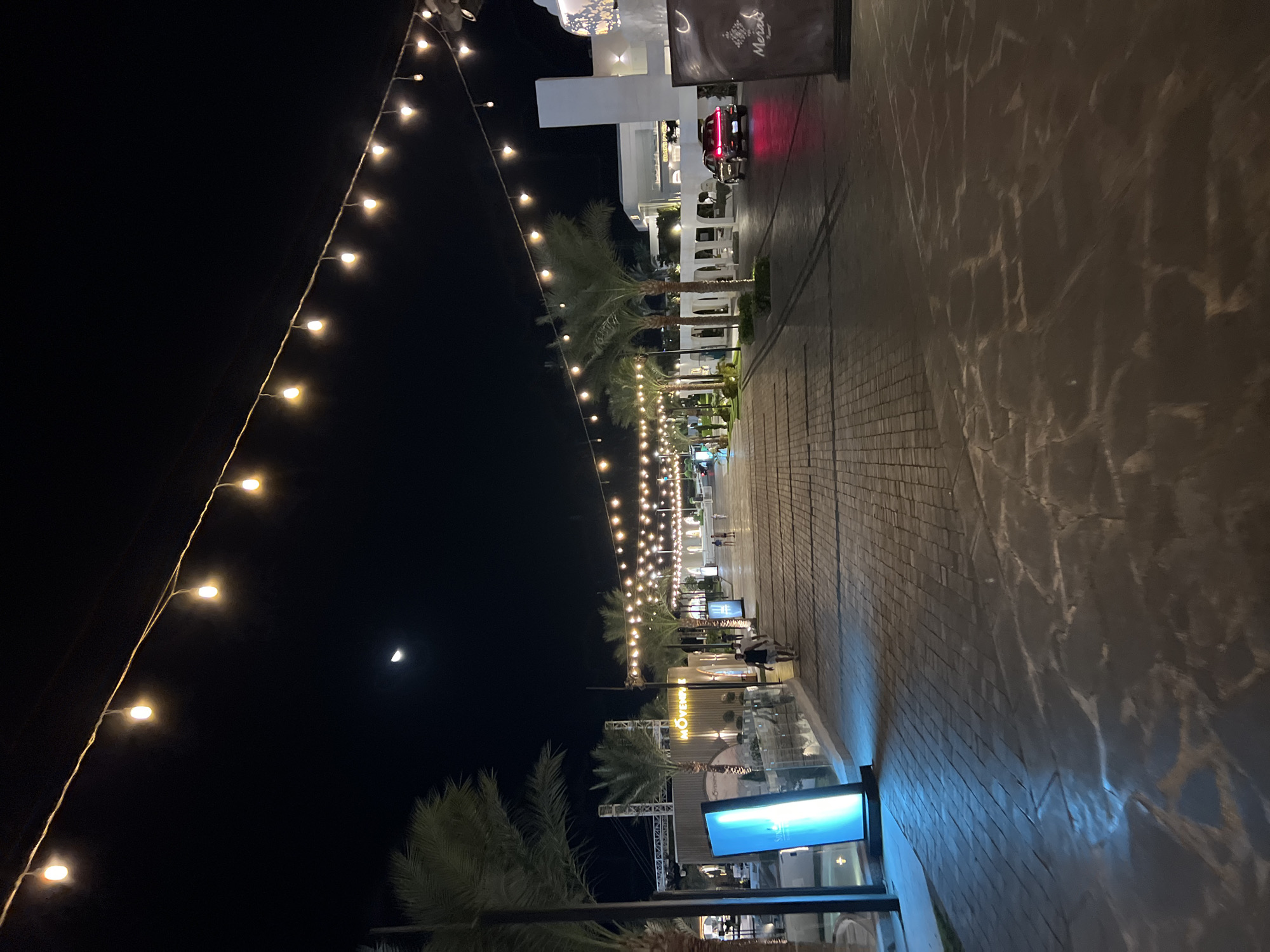 The image depicts a nighttime scene on a paved pedestrian walkway in Sharm El Sheikh, Egypt. String lights with visible bulbs are strung across the dark sky, illuminating the walkway which is paved with light-colored bricks. Palm trees line the sides of the walkway, with some exhibiting well-lit trunks. On the left, a vertical blue light display is visible, accompanied by a glass barrier. Further down the walkway, several people are walking. On the right, a white building with arched entrances is present, and a black SUV with illuminated red taillights is driving away. A sign with the word "Meraki" is also visible on the right side of the image. The moon is faintly visible in the upper left quadrant of the sky.