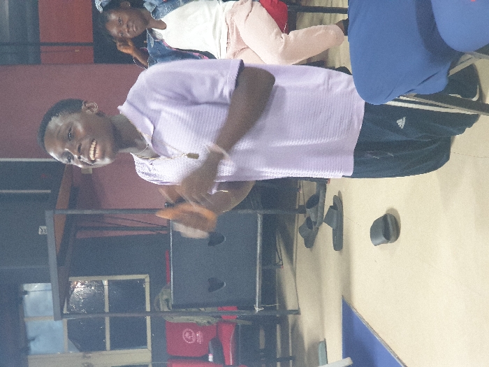A smiling young man in a light purple shirt and dark athletic pants stands indoors. He is looking at the camera with open hands. Behind him is a shelf-like structure, a red bucket, and a dark surface. In the background, a woman sits, and a blue object partially obscures the scene. Sandals and a dark object are on the floor.